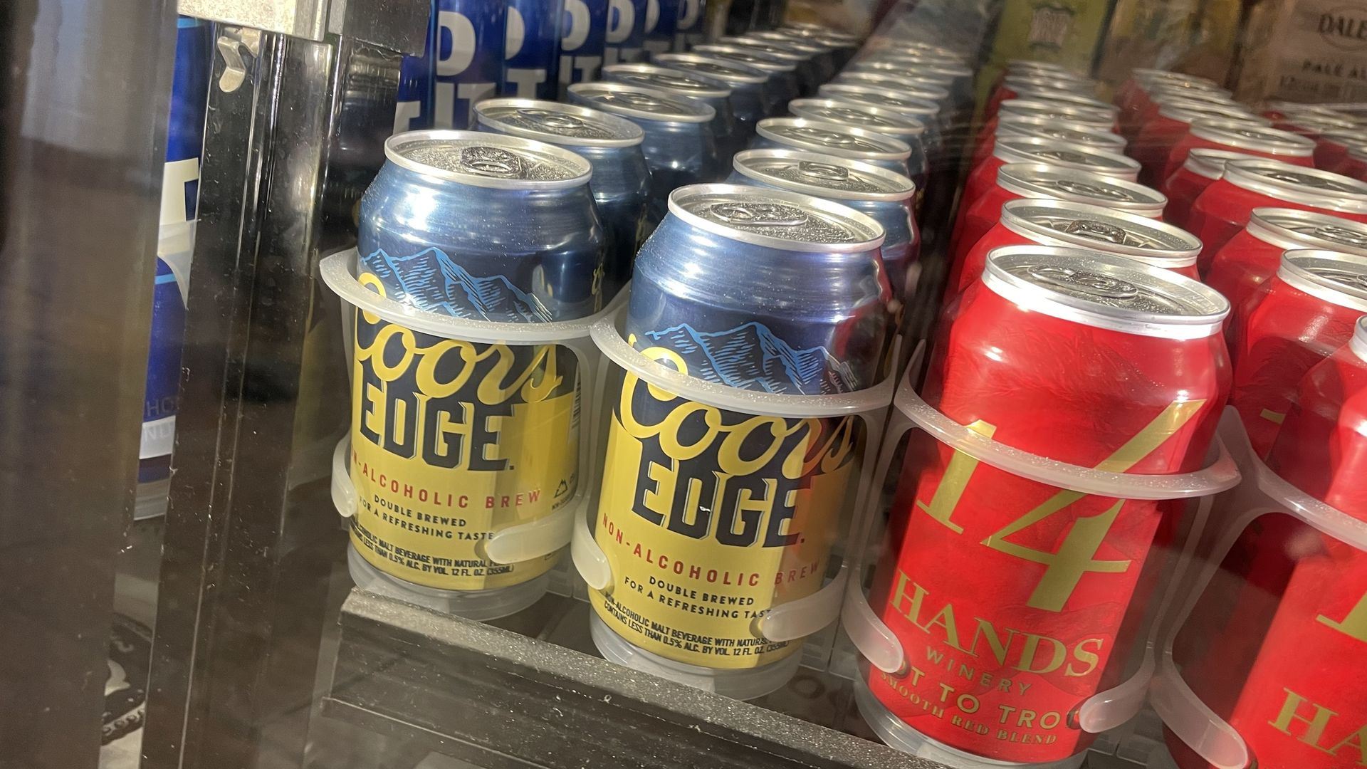 Four rows of canned beer bottles, with one reading COORS EDGE, in a blue and yellow can, and a bright red can with the words 14 HANDS on it. All the bottles are behind a glass refrigerator door. 