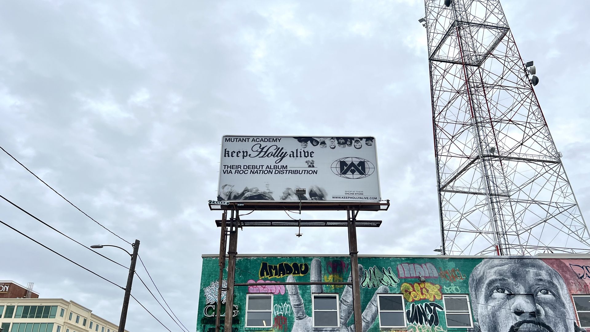 A pic of billboard that says "keep holly alive" next to a cell tower and a mural with gray skies in the background.