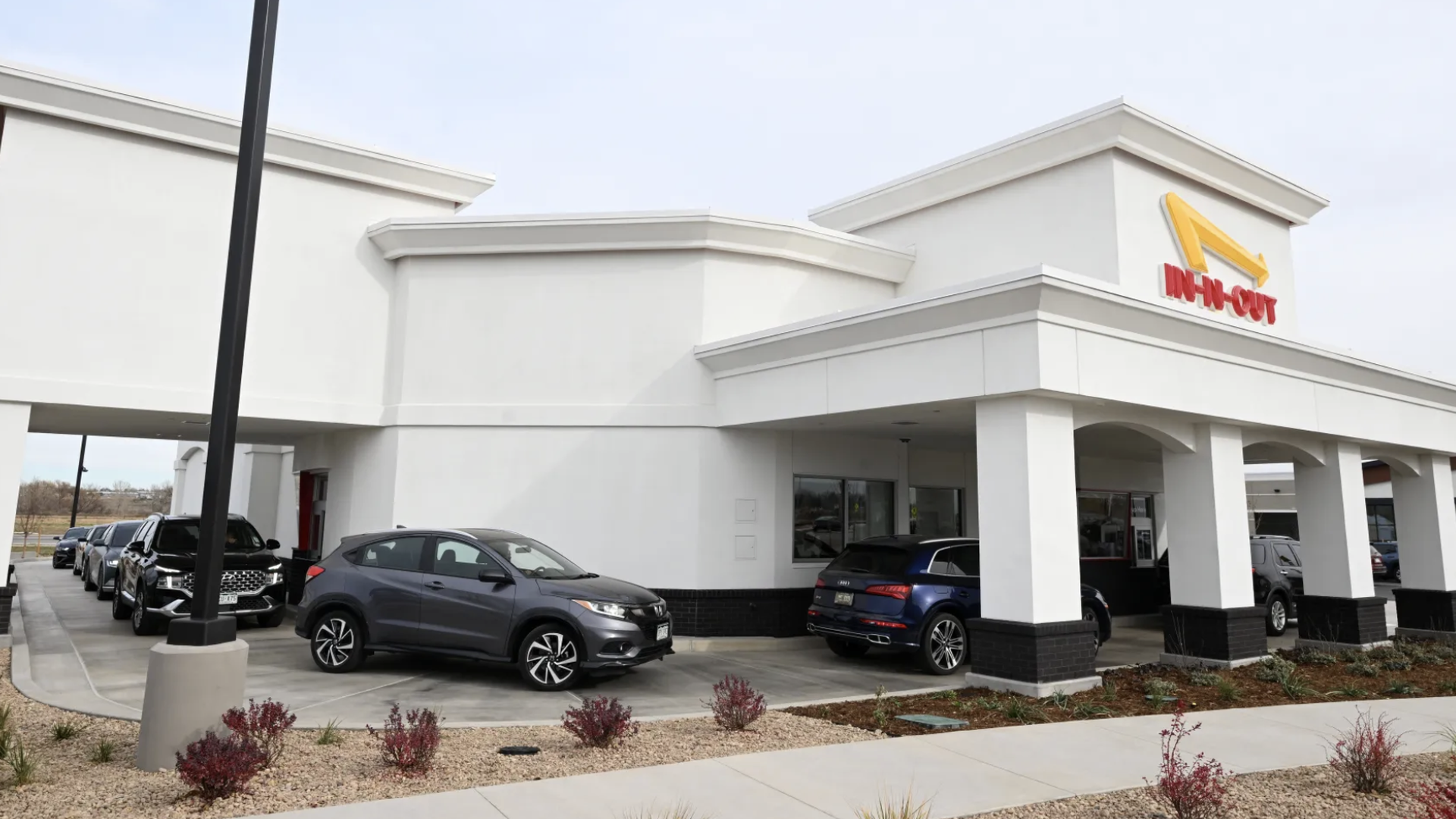 Longmont In-N-Out restaurant with line of cars in drive-thru lane, under cloudy sky with landscaped gravel and small bushes in front.