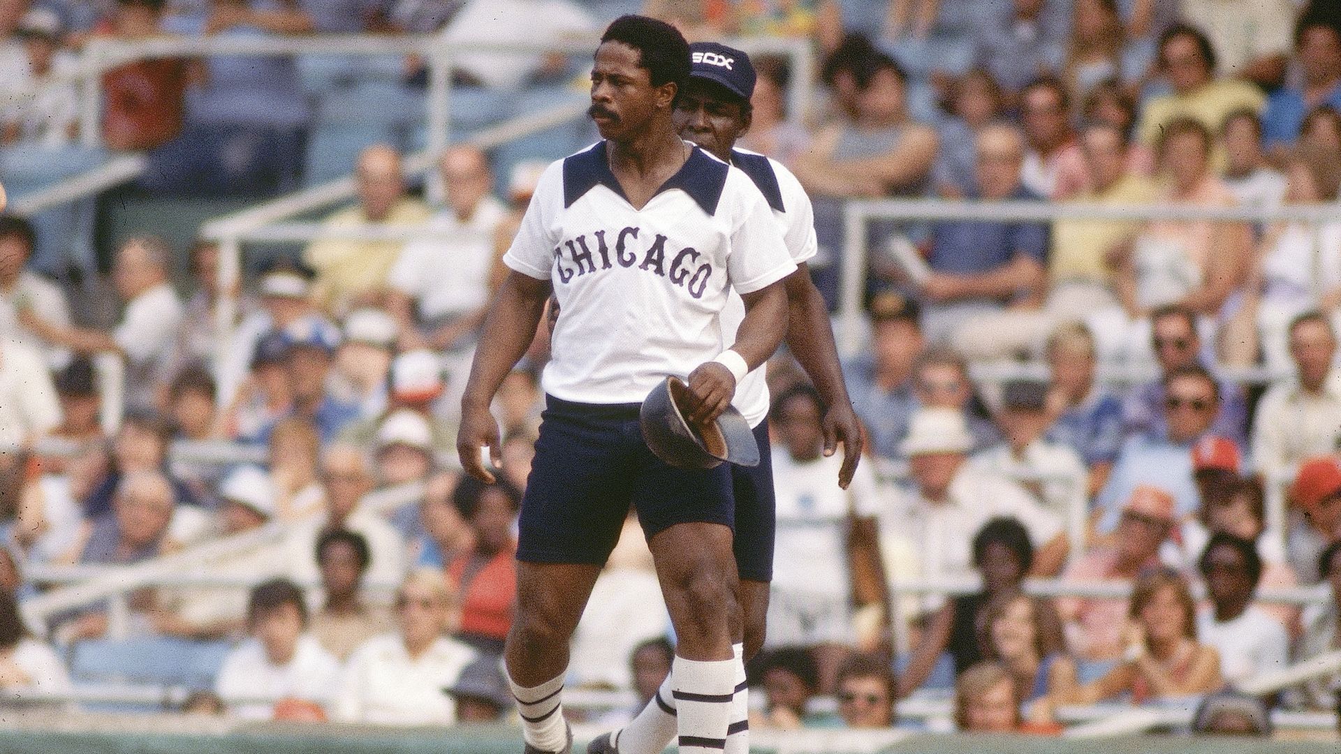 Two baseball players in vintage Chicago uniforms with white shirts and black shorts, one holding a black cap, on the field with a blurred crowd in the background.