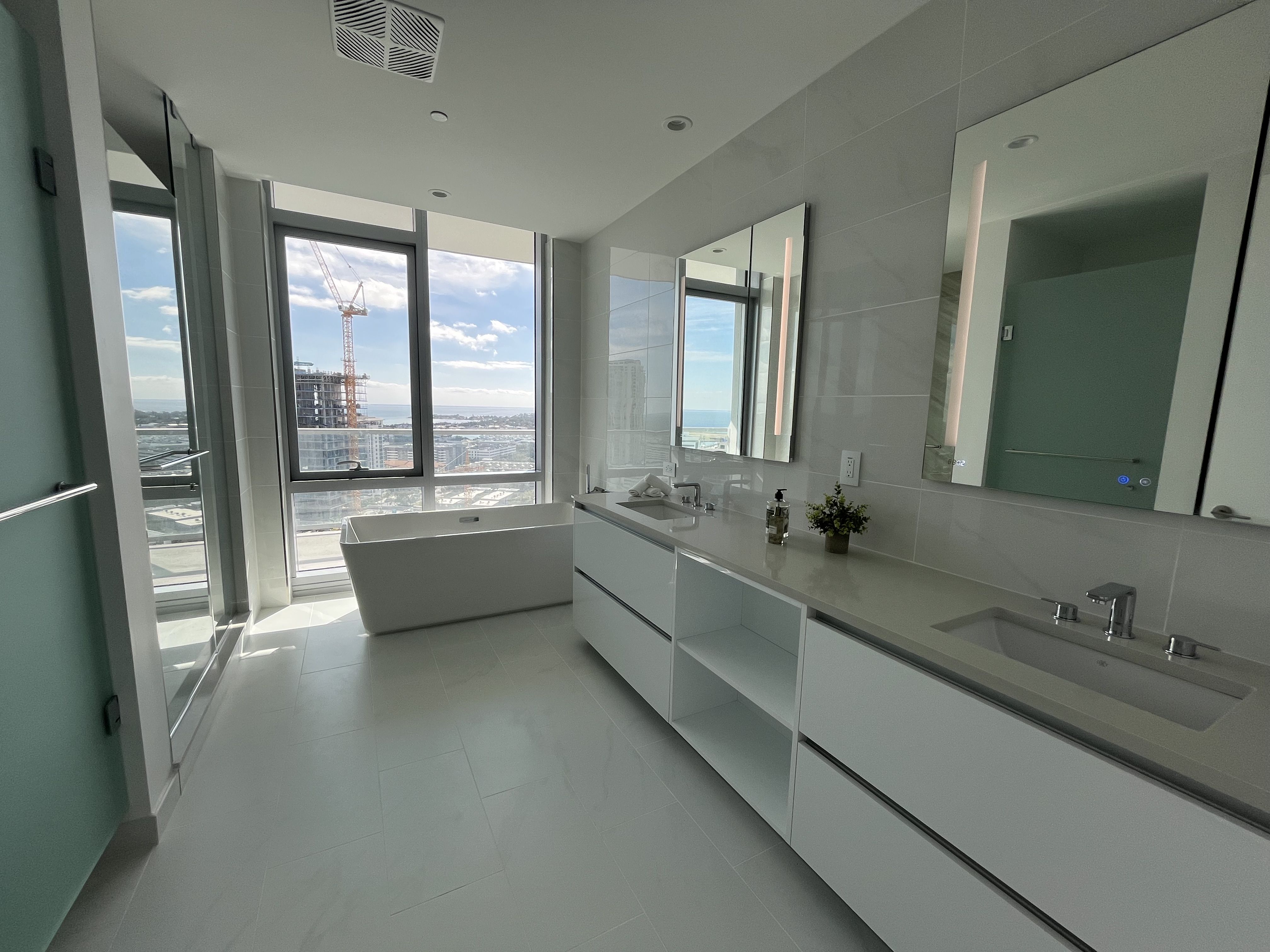Modern bathroom with white floor and walls, double sink vanity with mirrors, freestanding white bathtub by large window overlooking city and ocean, and green accent wall