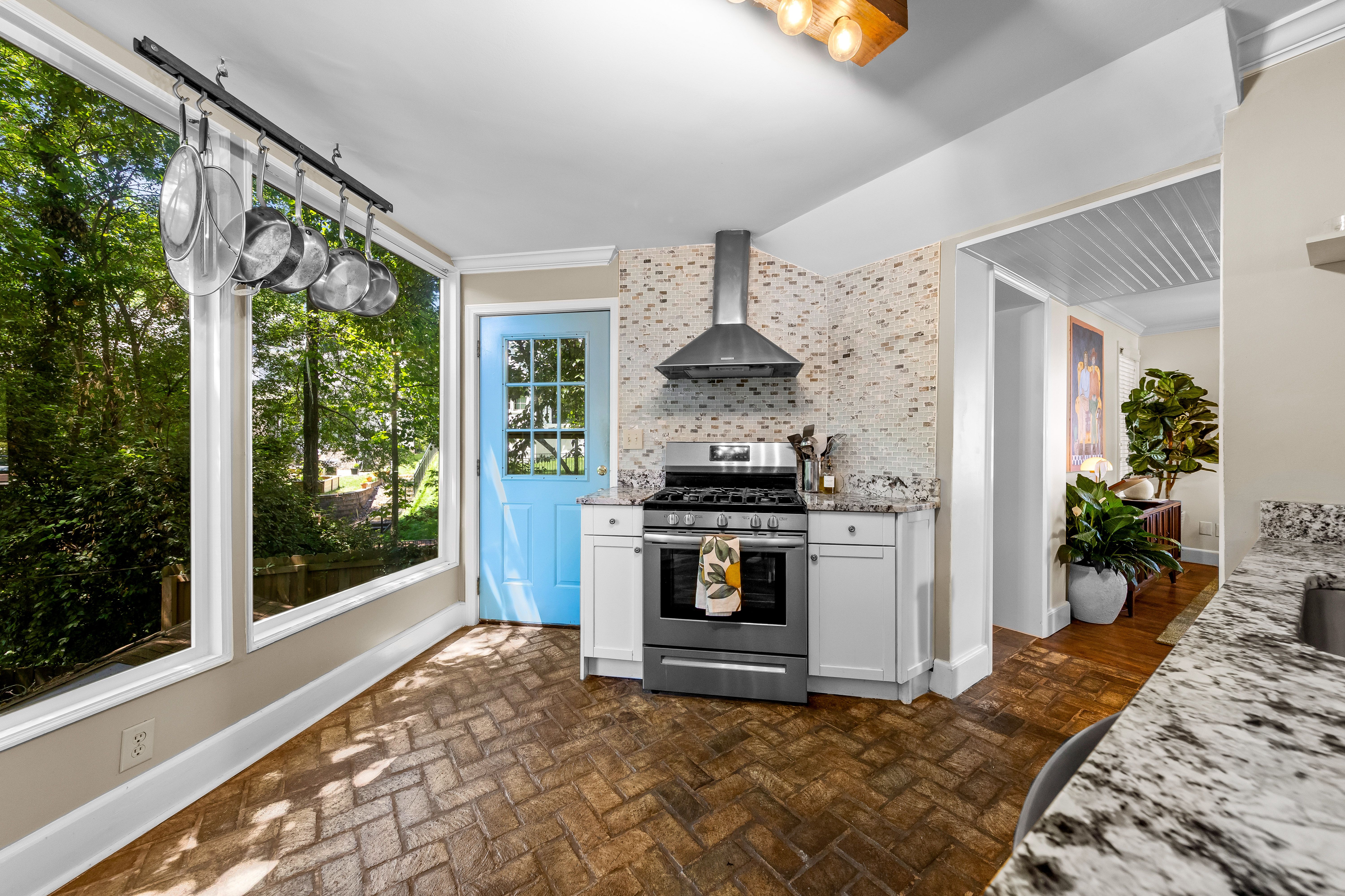 Bright kitchen with a row of stainless pots hanging from a ceiling pot rack, large windows showing greenery, a blue door, a stainless gas range with mosaic tile backsplash, and granite countertops.