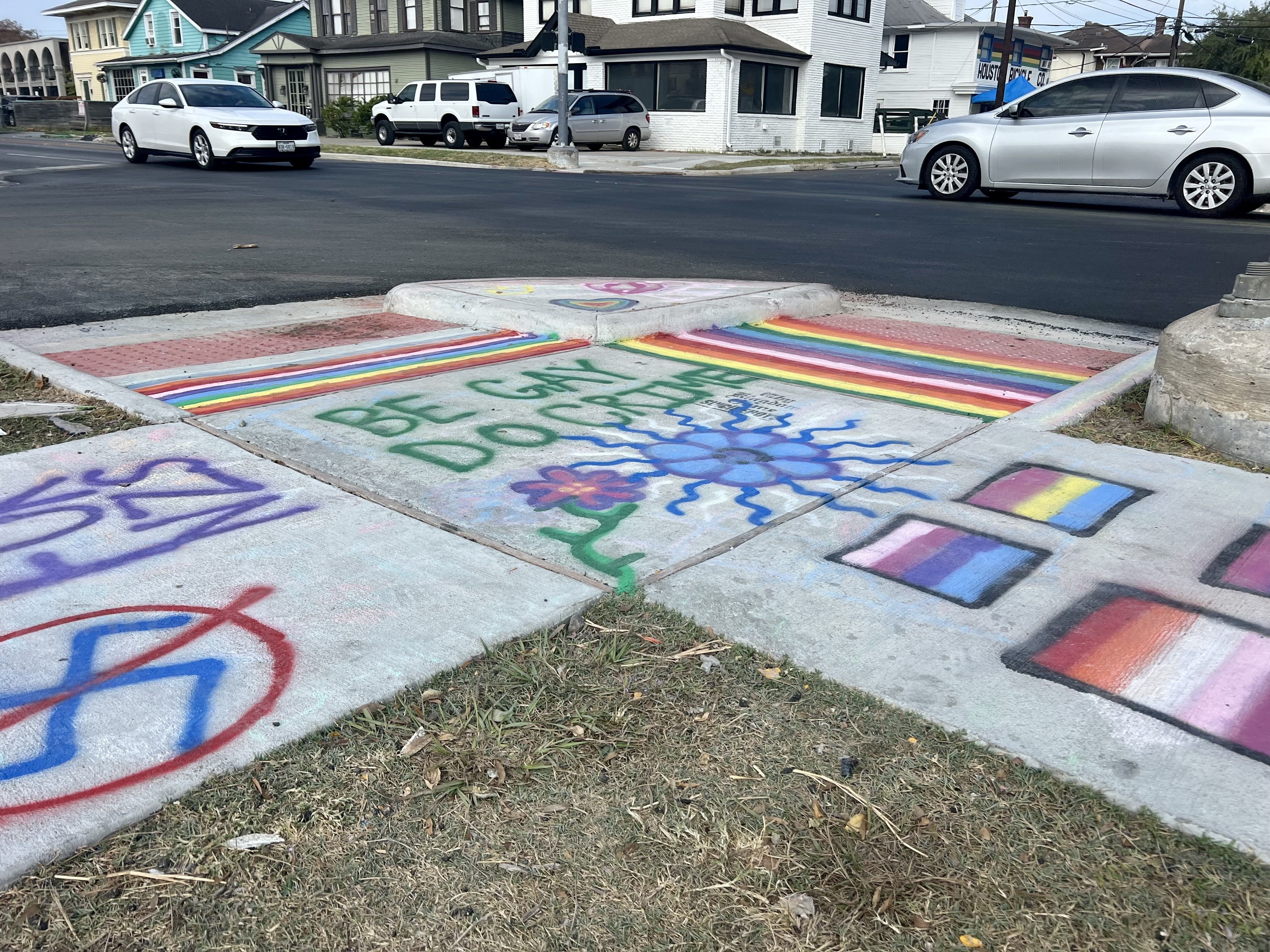 Sidewalk corner with colorful rainbow stripes and various LGBTQ+ pride flags, green spray-painted words "BE GAY DO CRIME," and a blue sun and a flower graffiti art.