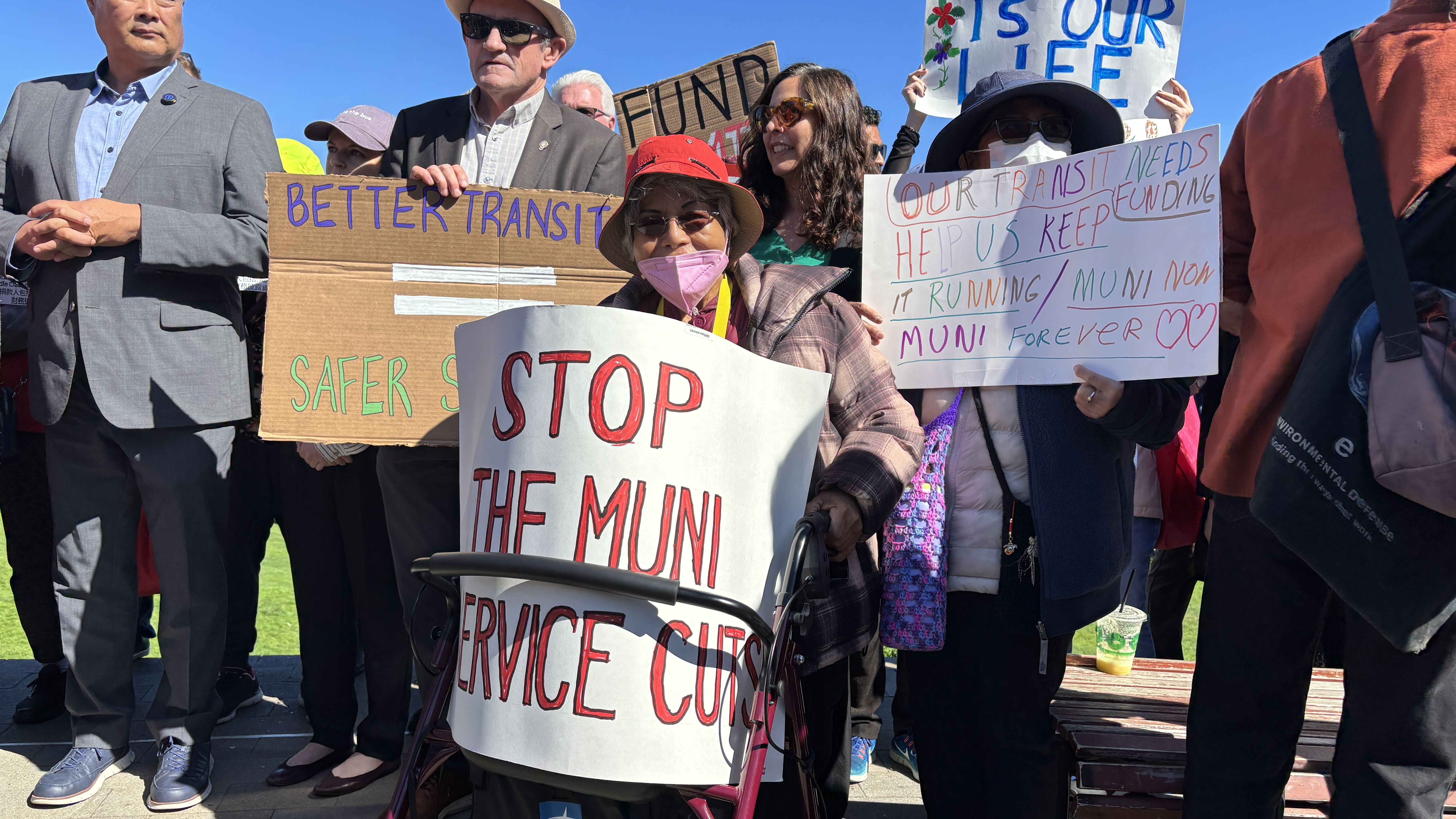 Group of people at a daytime protest holding signs about transit funding and service, including "STOP THE MUNI SERVICE CUTS" and "OUR TRANSIT NEEDS HELP US KEEP IT RUNNING/MUNI FOREVER."