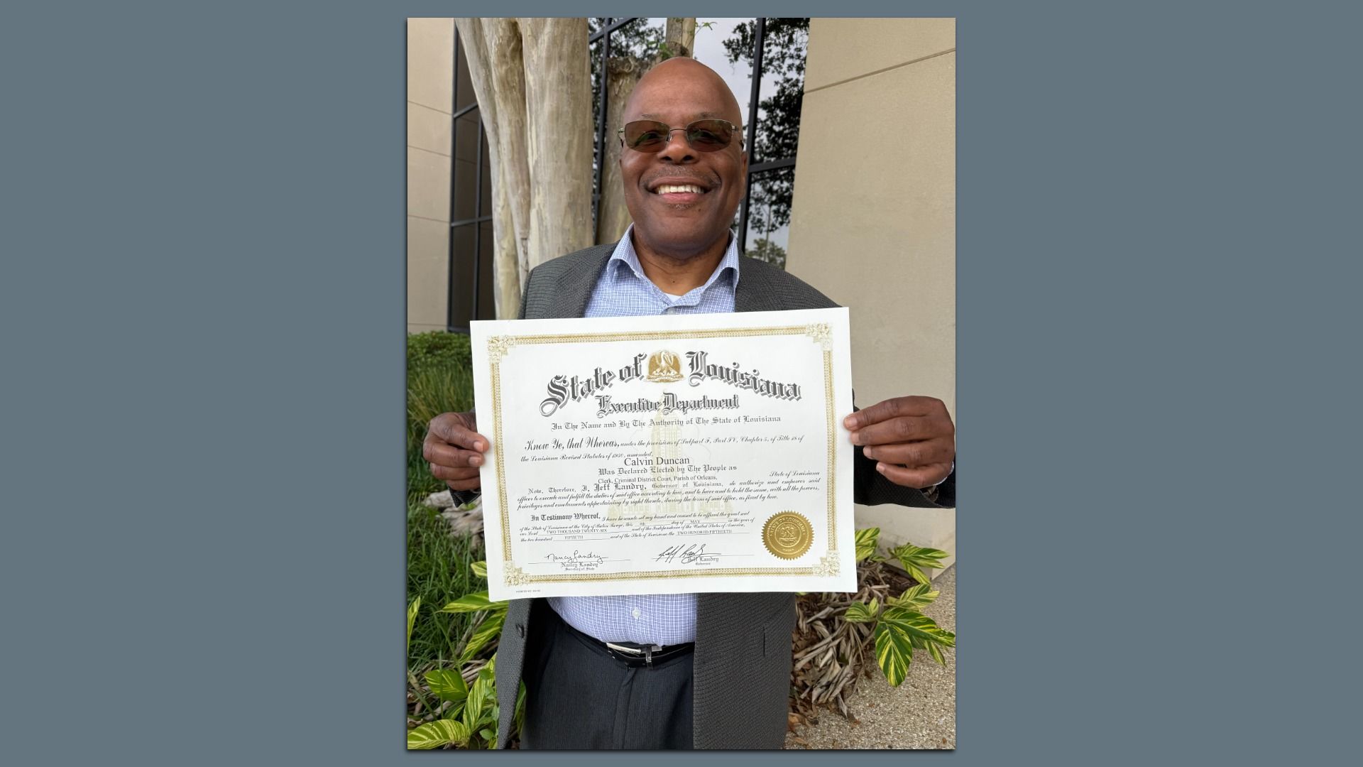 Smiling man in a gray suit and light blue shirt, wearing sunglasses, holds a large ornate certificate outdoors beside trees and a beige wall.