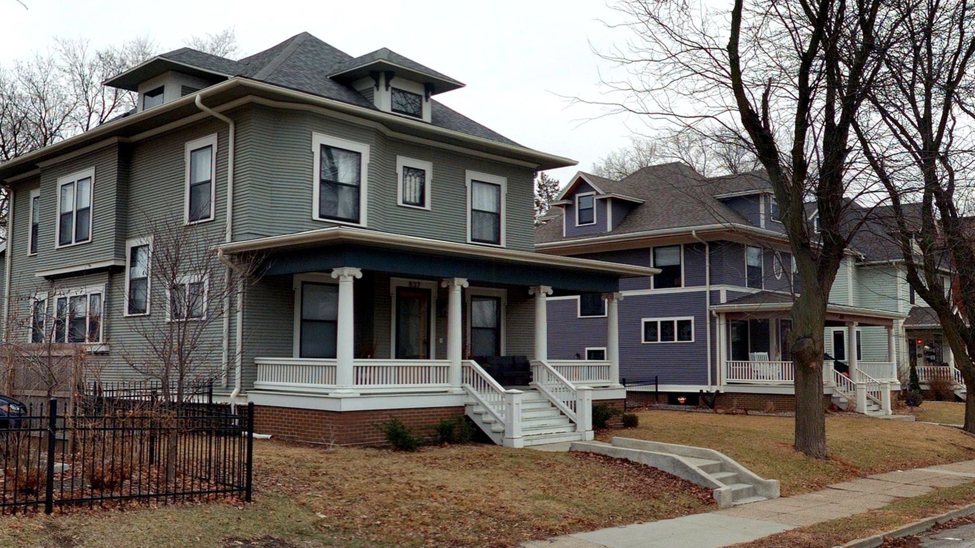 Row of two-story houses with wraparound porches and white railings on a quiet residential street. Gray and blue siding, brick foundations, leafless trees, and a street lamp nearby.