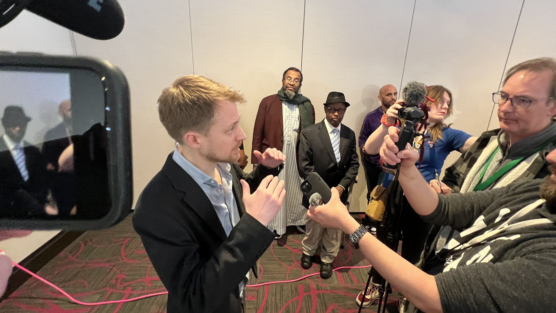 A white man in a suit jacket and blue button-down shirt gestures in front of a group of people holding up recording equipment, cameras and phones.