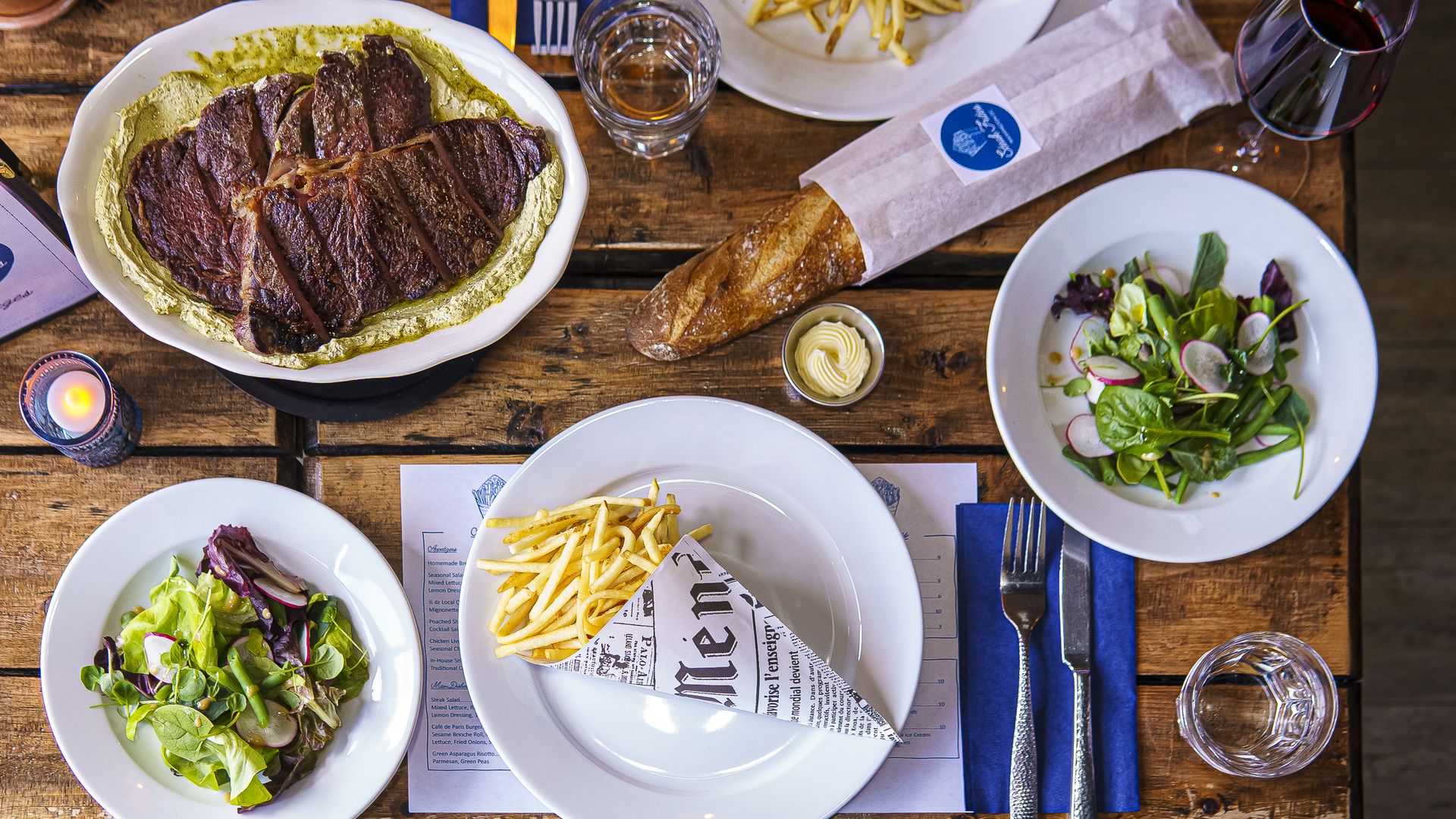 A wooden table with white plates of salad, French fries, a baguette and steak 