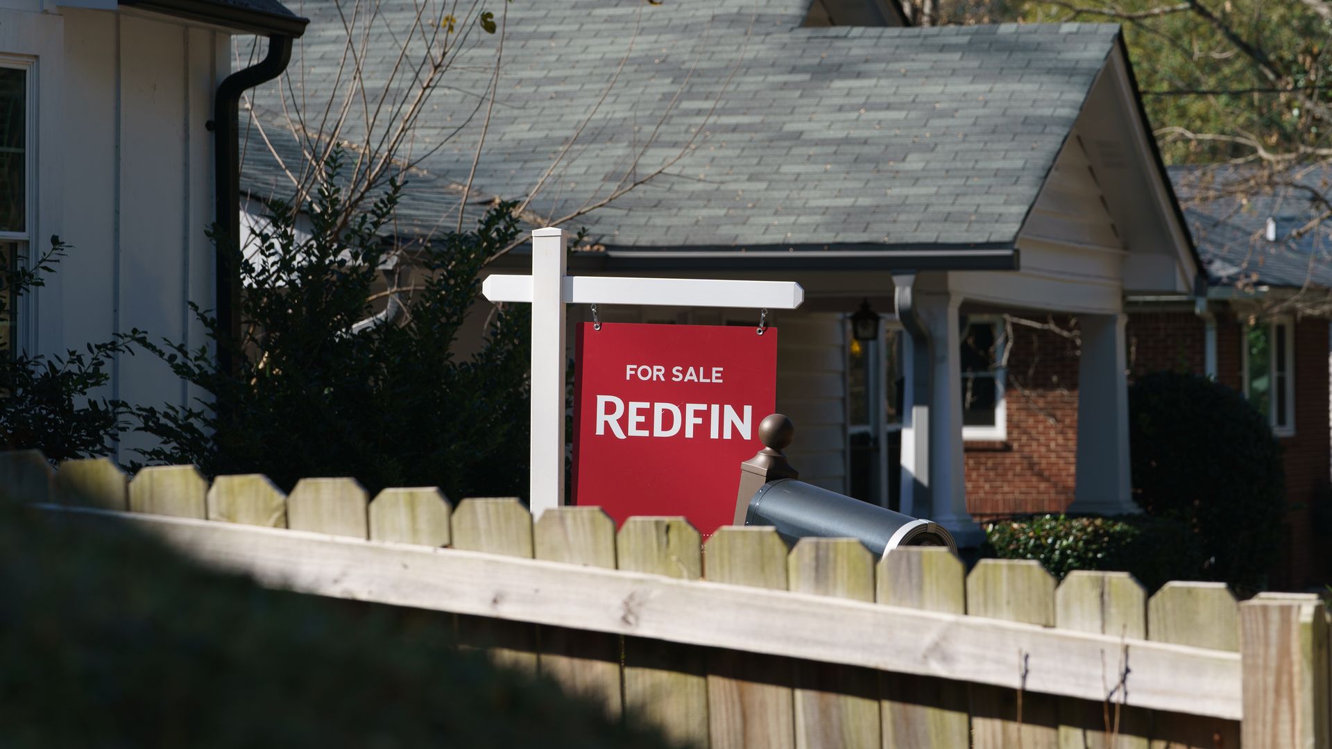 A for-sale sign behind a fence in the yard of a white, one-story house.