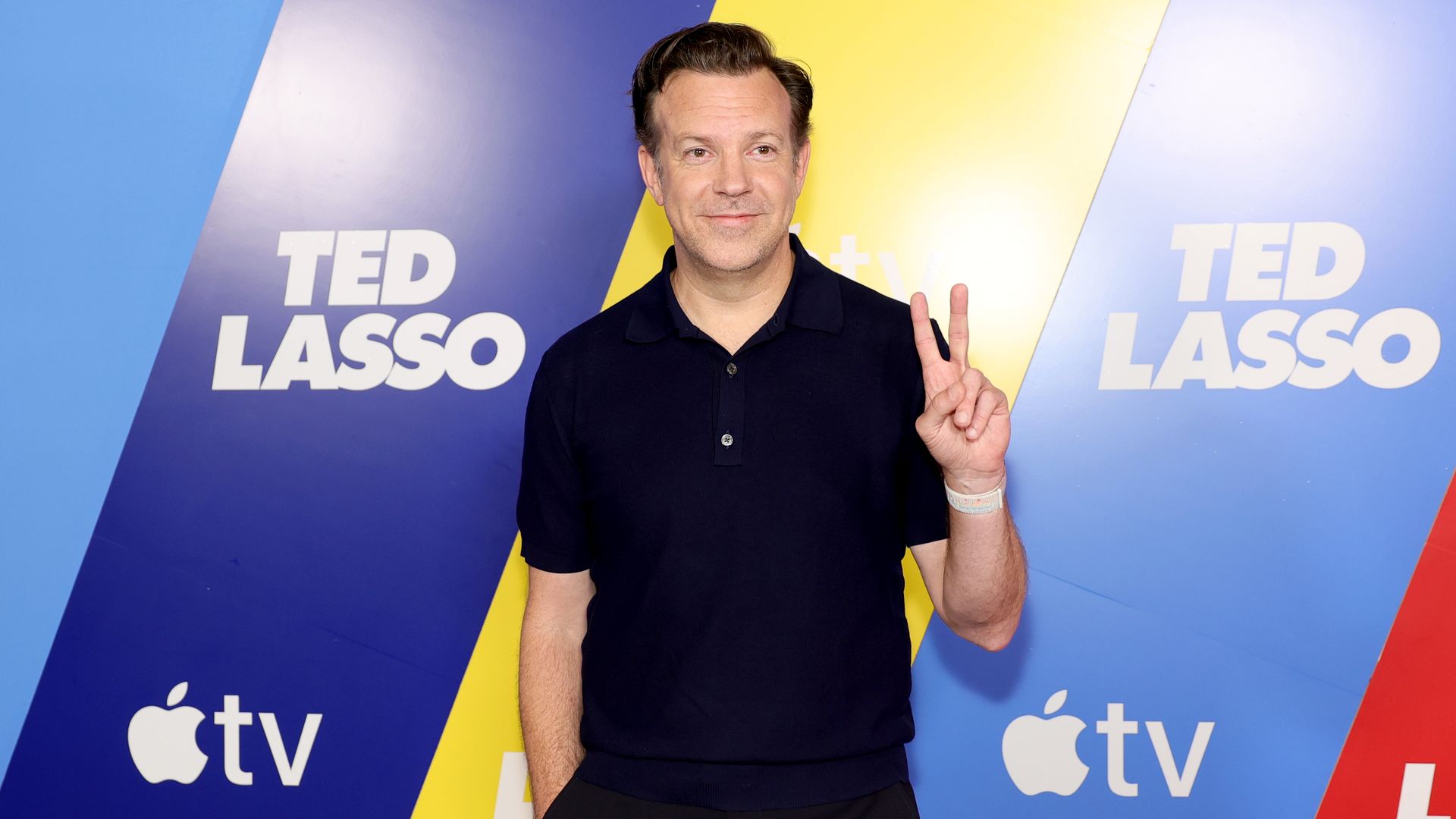 PErson in a dark polo poses in front of a blue, yellow, and purple backdrop with the text "TED LASSO" and Apple TV logos, smiling and flashing a peace sign.