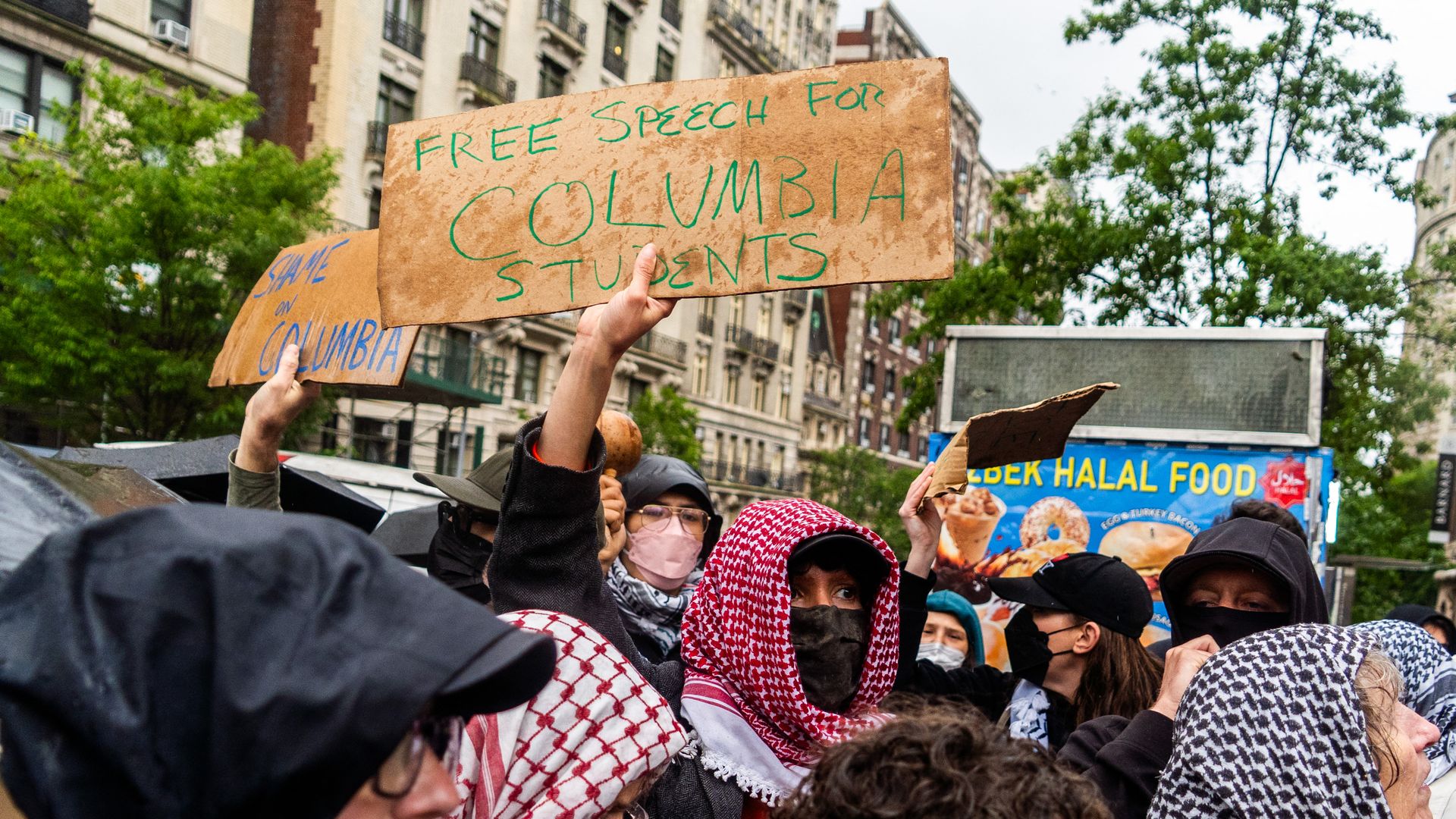Crowd of masked protesters in hoodies and keffiyeh scarves holding cardboard signs including one that reads \