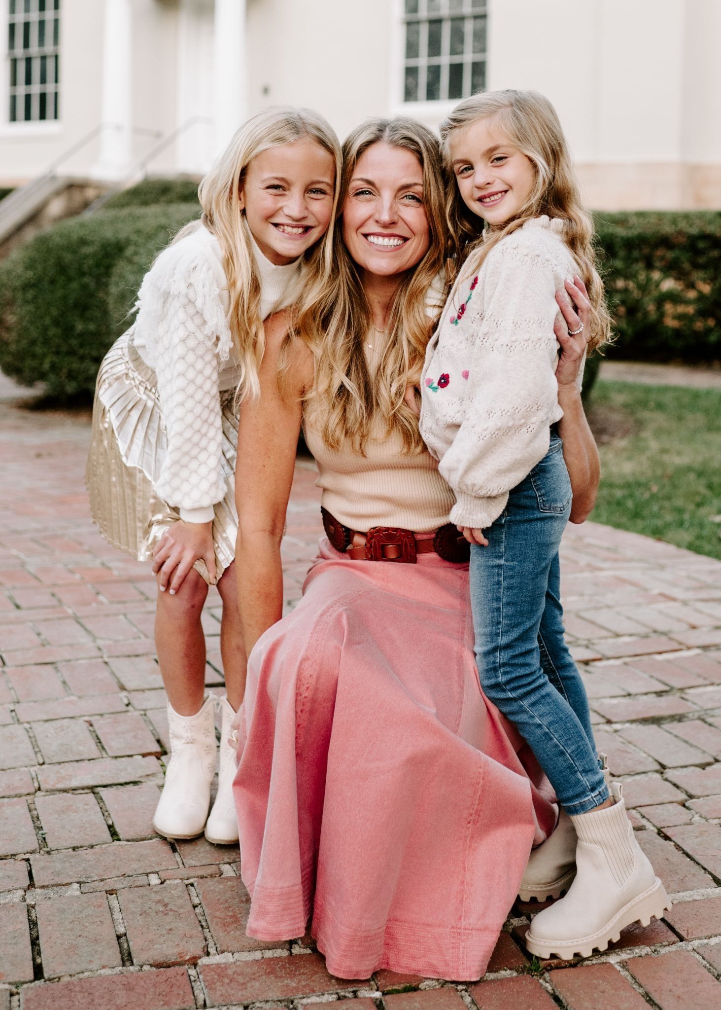 Smiling woman with long blonde hair kneels between two blonde girls outdoors; woman wears a pink skirt and beige top, left girl wears white sweater and gold skirt, right girl wears cream sweater and blue jeans.