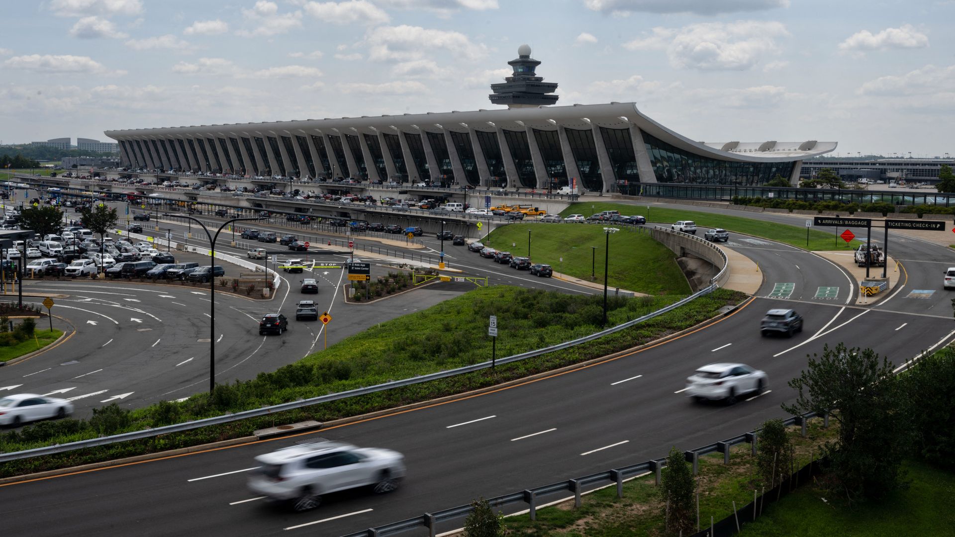 Wide shot of a modern airport terminal with a tiered control tower in the center, a long curved white roof, glass walls, and a busy roadway with cars in the foreground under a cloudy sky.