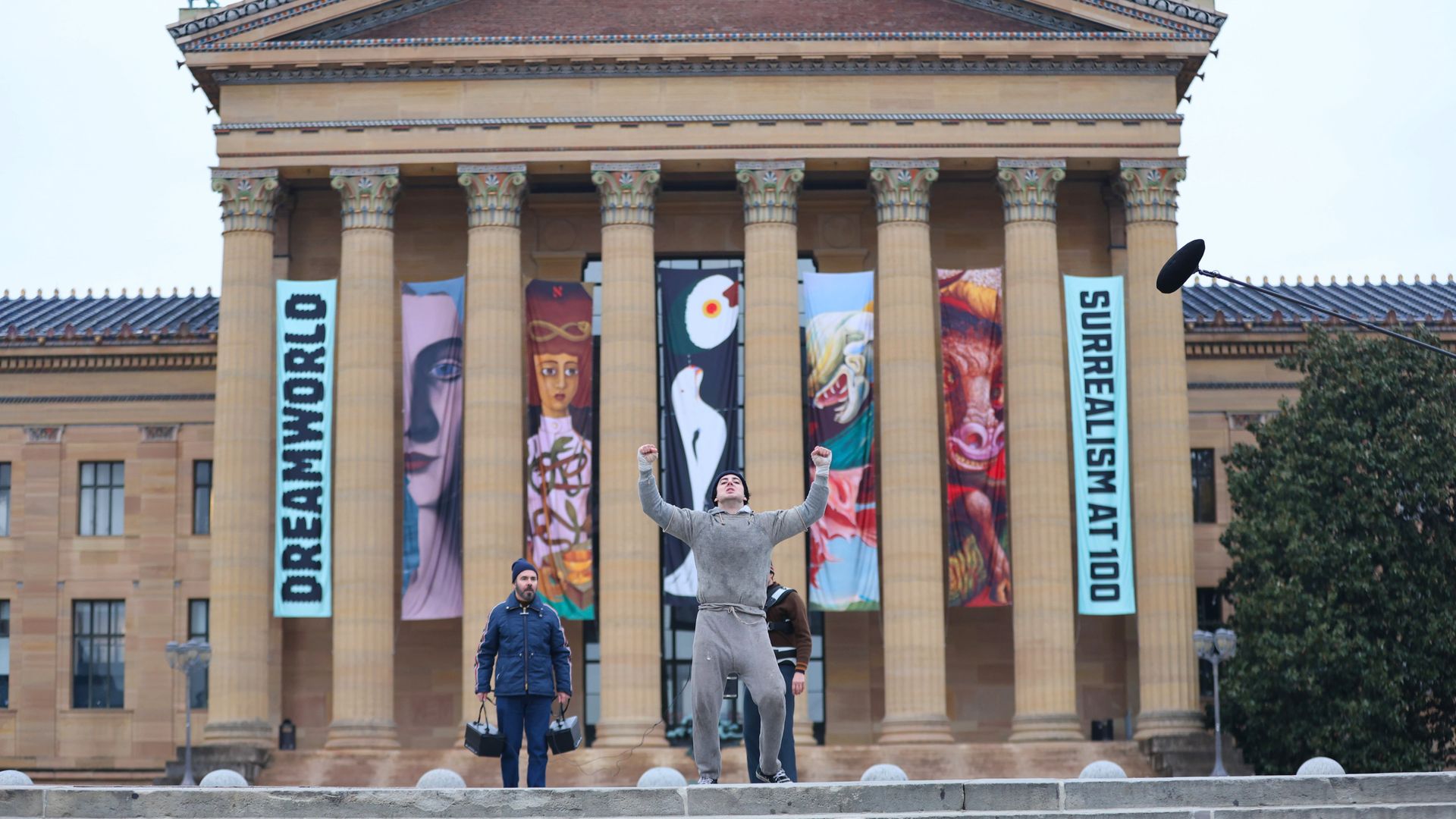 A man in a gray sweatshirt and sweatpants and beanie holding his hands up in front of a neo-classical building with several columns. 