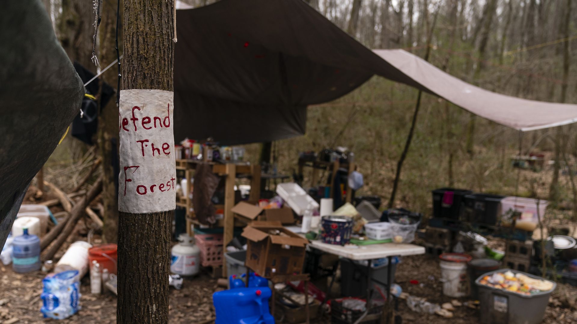 A photo of abandoned campsite under a tarp in the woods occupied by activists opposed to a public safety training center and a tree with a note that says "Defend the forest"