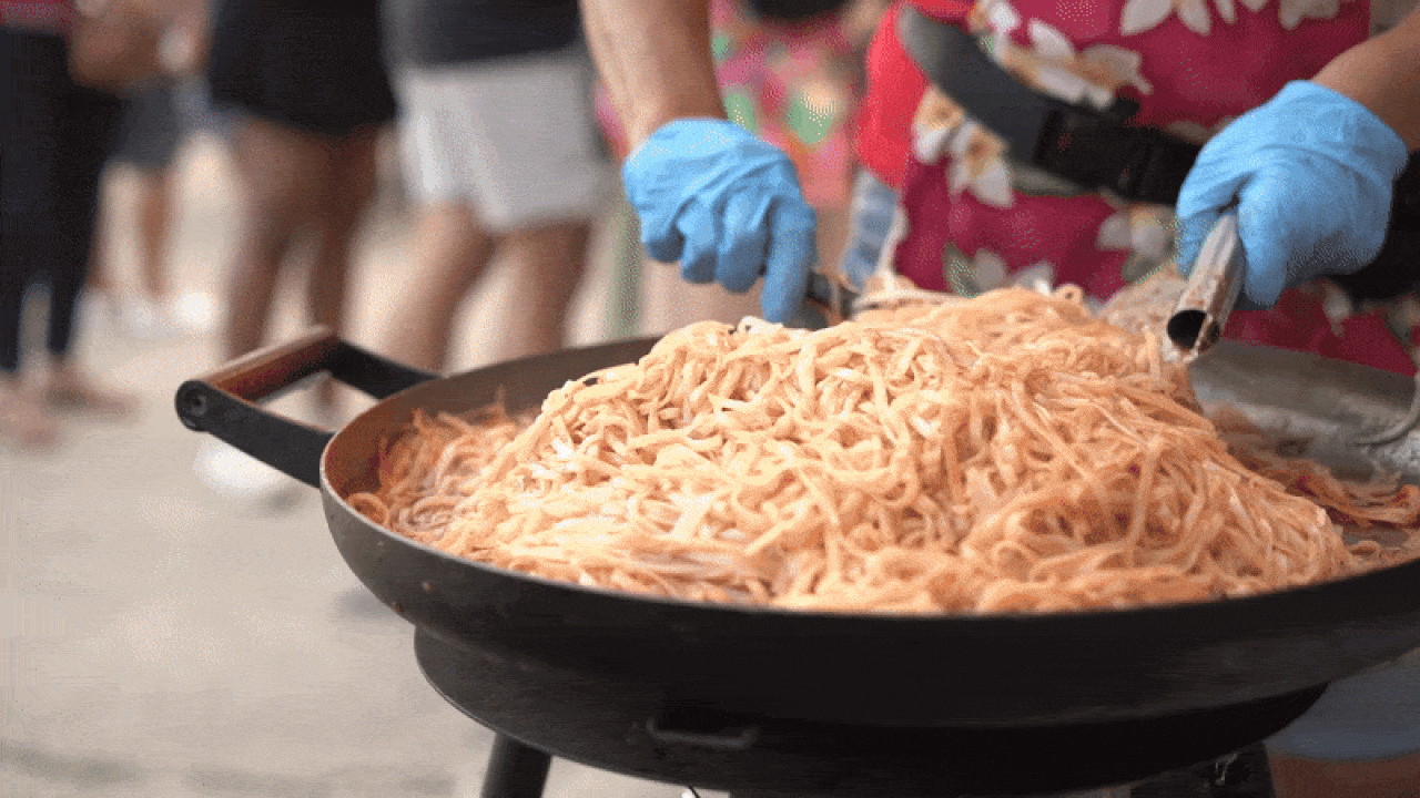 Person wearing blue gloves and a floral red apron cooking or stirring a large pan of orange-colored noodles outdoors, with blurred people standing in the background.