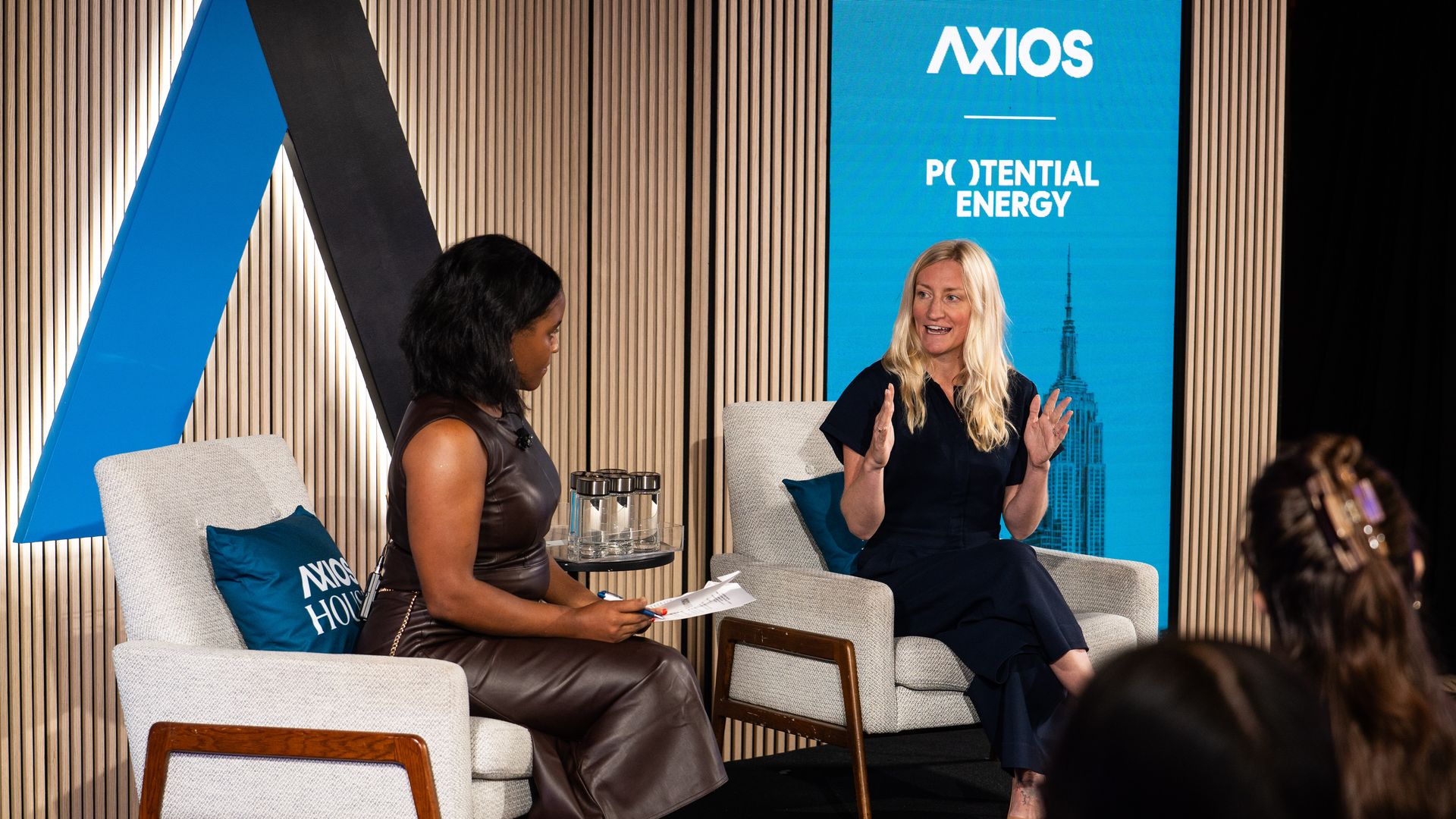 Two women seated in armchairs on a stage at an Axios event, one in a brown leather dress and the other in a navy outfit, engaged in a discussion.