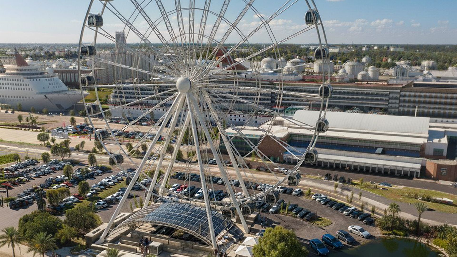 Large ferris wheel with black gondolas near parking lots and a pond, cruise ships docked in background under blue sky with scattered clouds, palm trees lining the area.