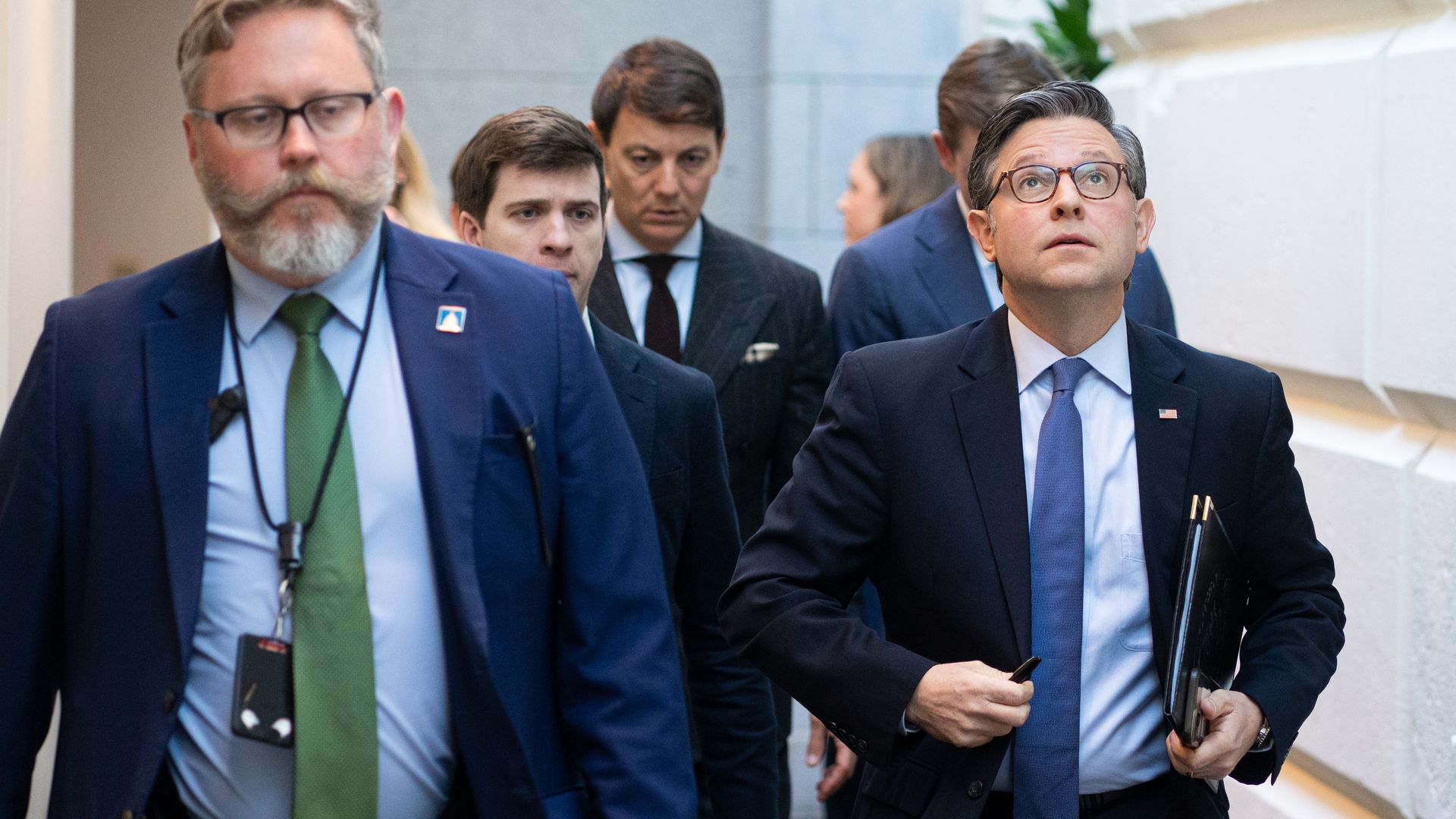 Group of formally dressed men walking in a corridor, one in front left wearing green tie and glasses, another on right with blue tie and glasses looking upward, neutral background.