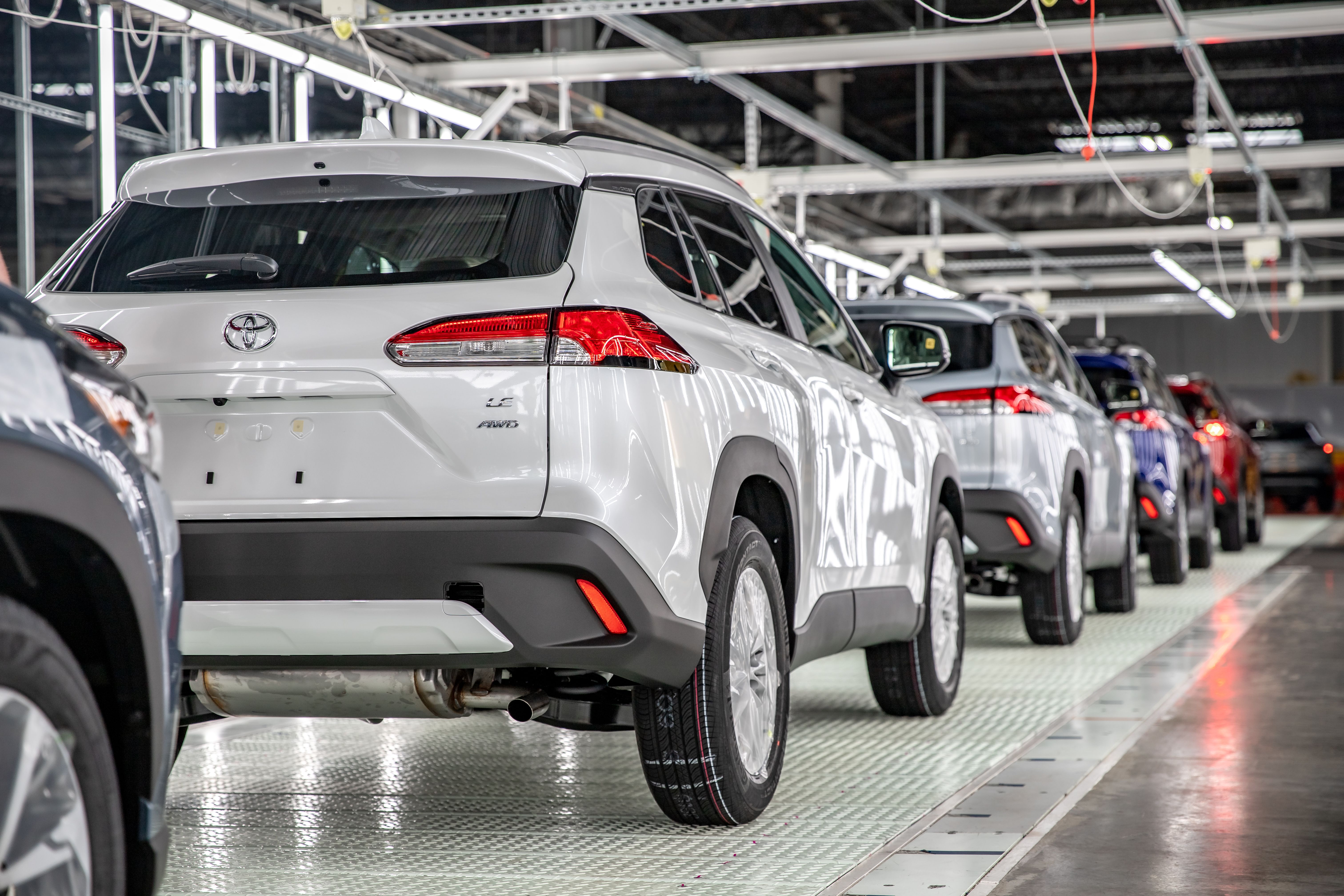 White Toyota SUVs lined up on a car assembly line inside a factory, seen from the rear sides; overhead metal framework and wiring loom above as other vehicles extend into the background.