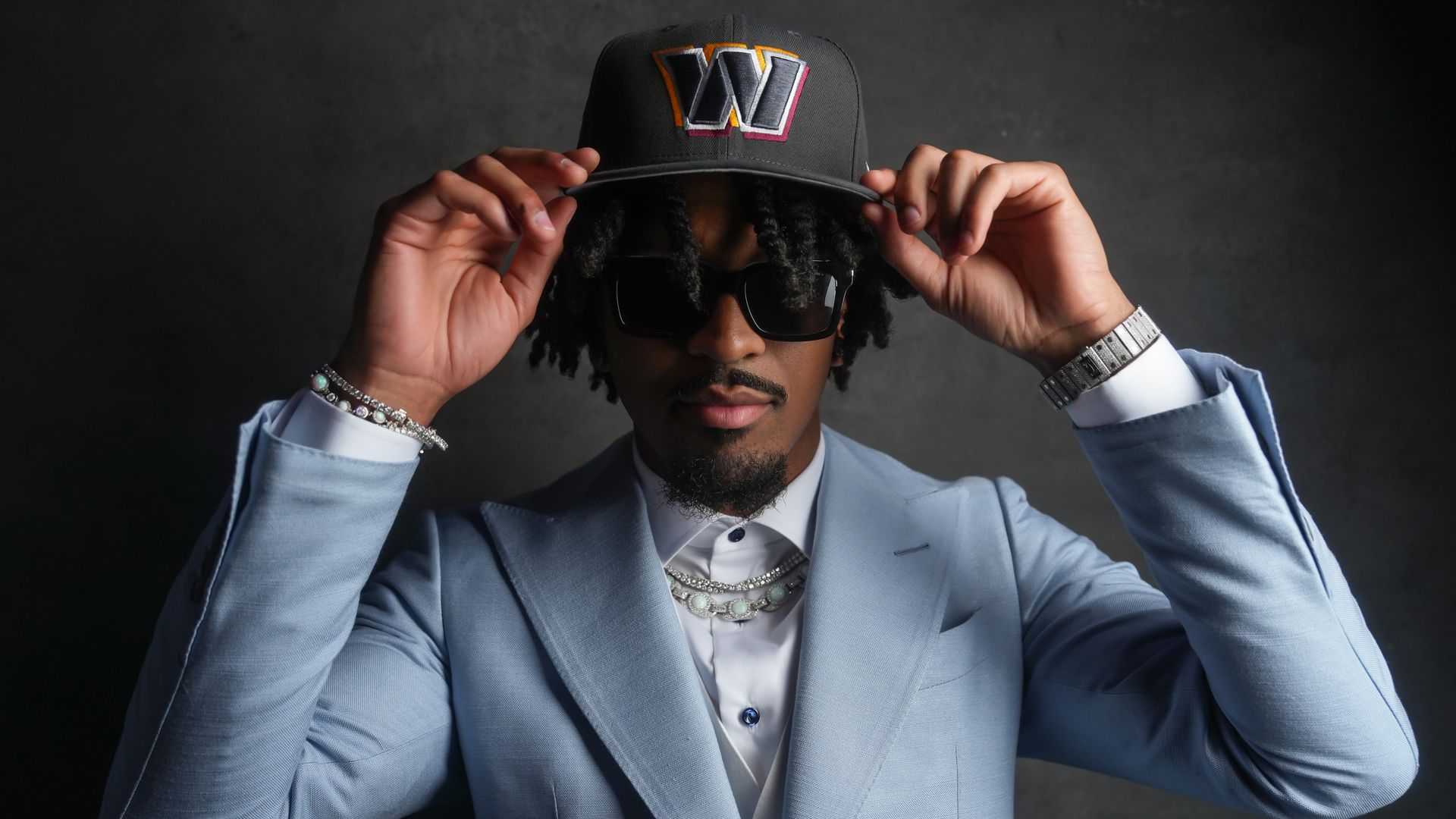 Jayden Daniels places a Washington Commanders black hat on his head, while wearing sunglasses and a light blue suit.