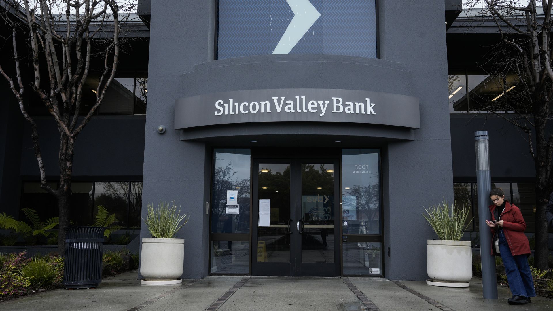 The entrance to Silicon Valley Bank's California headquarters, a dark gray building with two leafless trees and a double glass door.