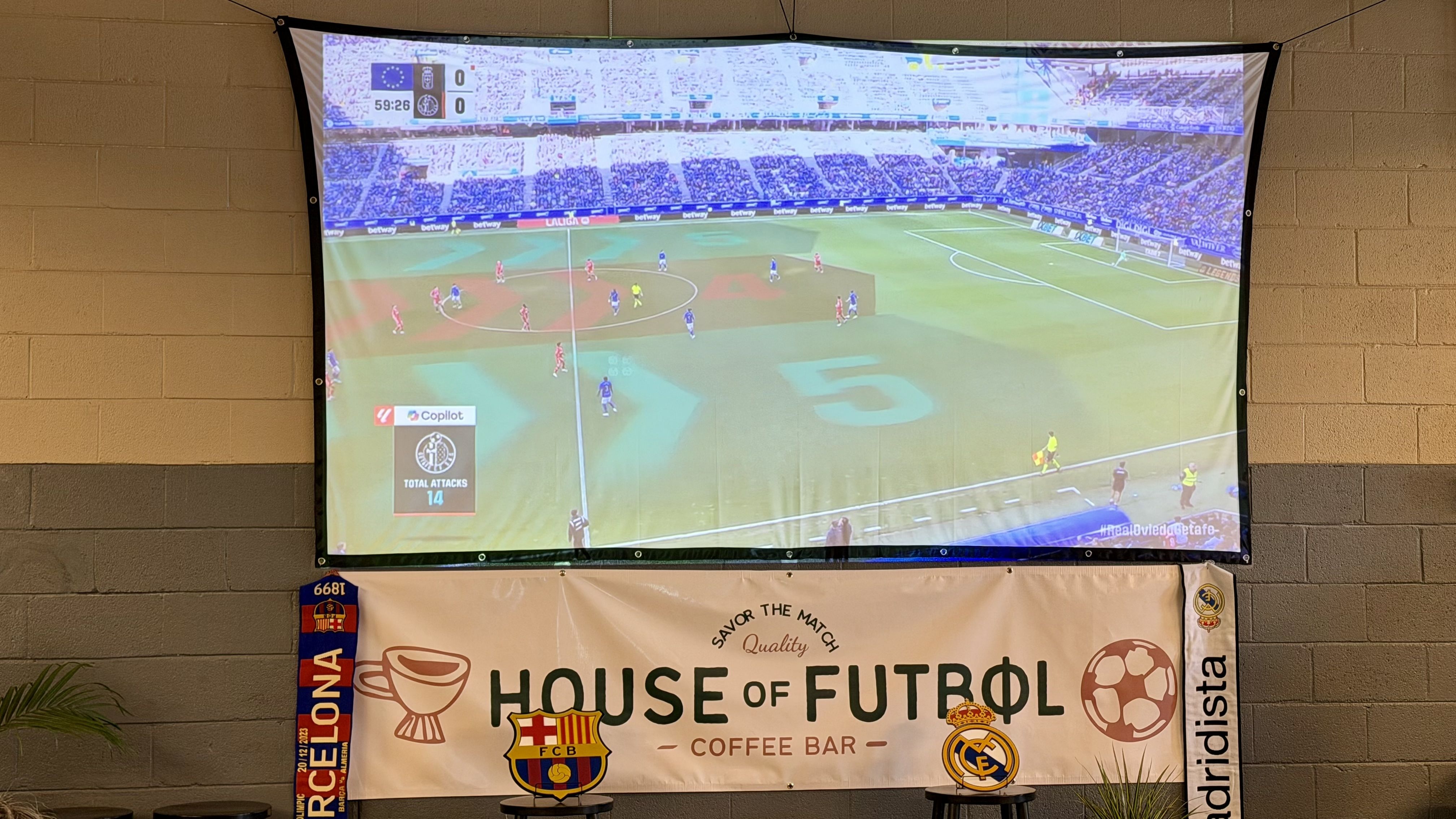Indoor wall-mounted screen showing a soccer match: players in red and blue on a green pitch, a full stadium in the background; a large "House of Football" banner hangs below.