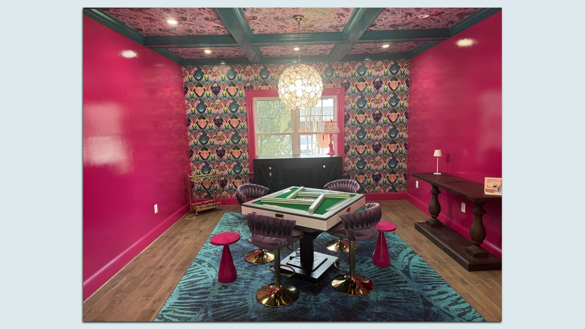 Bright pink-walled mahjong room with bold patterned back wall, green ceiling beams, a round chandelier, a green square table, four purple tufted chairs, pink stools, and a dark wood console by a window.