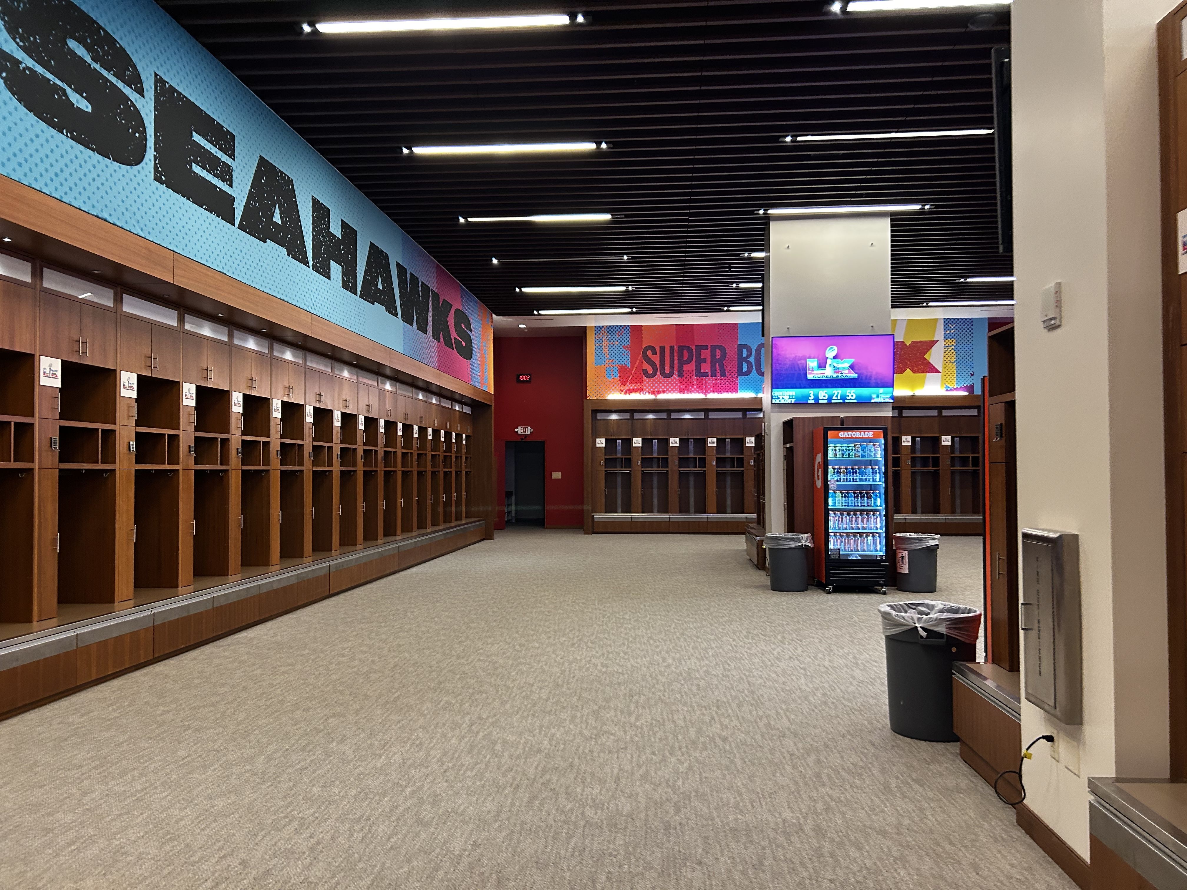 Empty Seattle Seahawks locker room with wooden lockers along walls, large blue and red "SEAHAWKS" and "SUPER BOWL" banners above, a Gatorade fridge, and a TV screen.