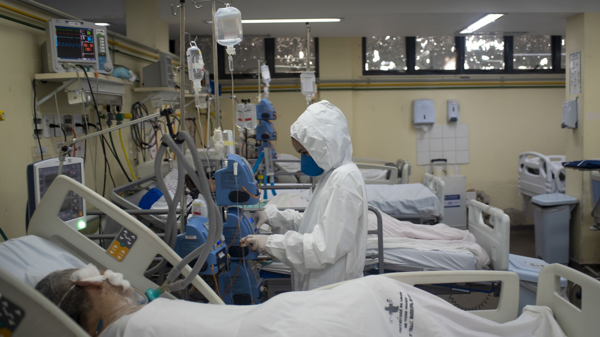 A hosptial worker in a white hooded hazmat suit stands among hospital beds where patients appear to be on ventilators.