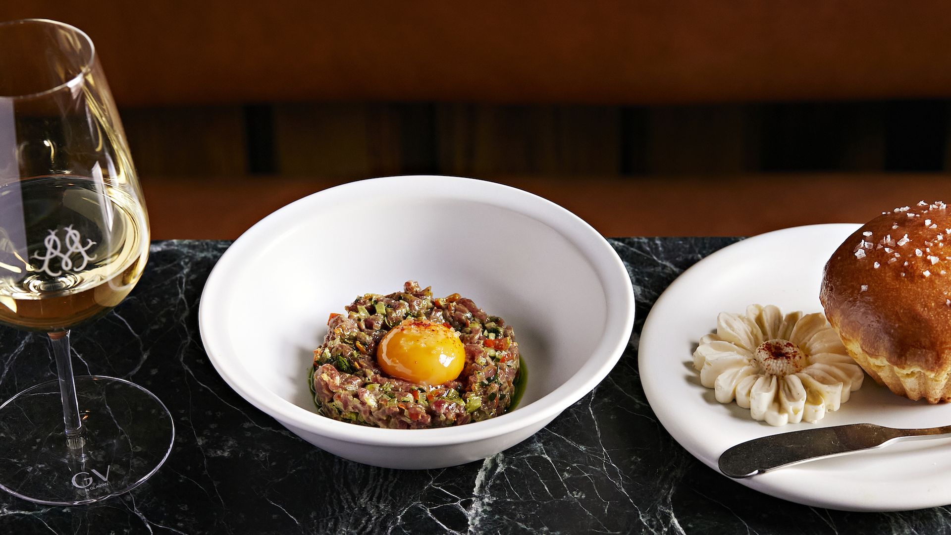 A glass of white wine next to a bowl of steak tartare topped with a raw egg yolk, and a plate with a salted brioche bun and a flower-shaped butter pat on a dark marble table.