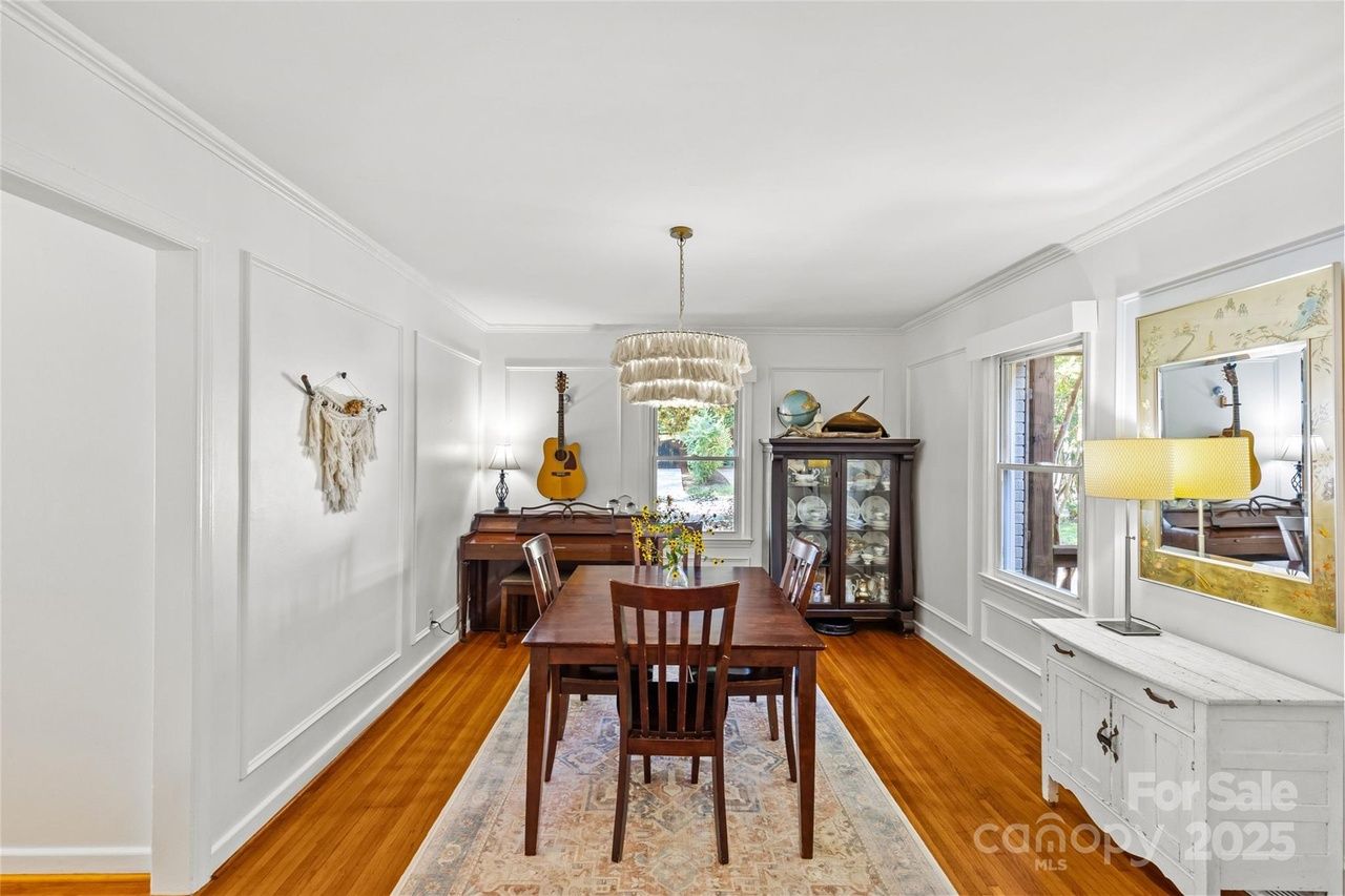 Bright dining room with white walls, wood floor, dark wooden table and chairs, a guitar above a piano, and a glass cabinet displaying dishes. A white sideboard with a lamp and mirror is on the right.