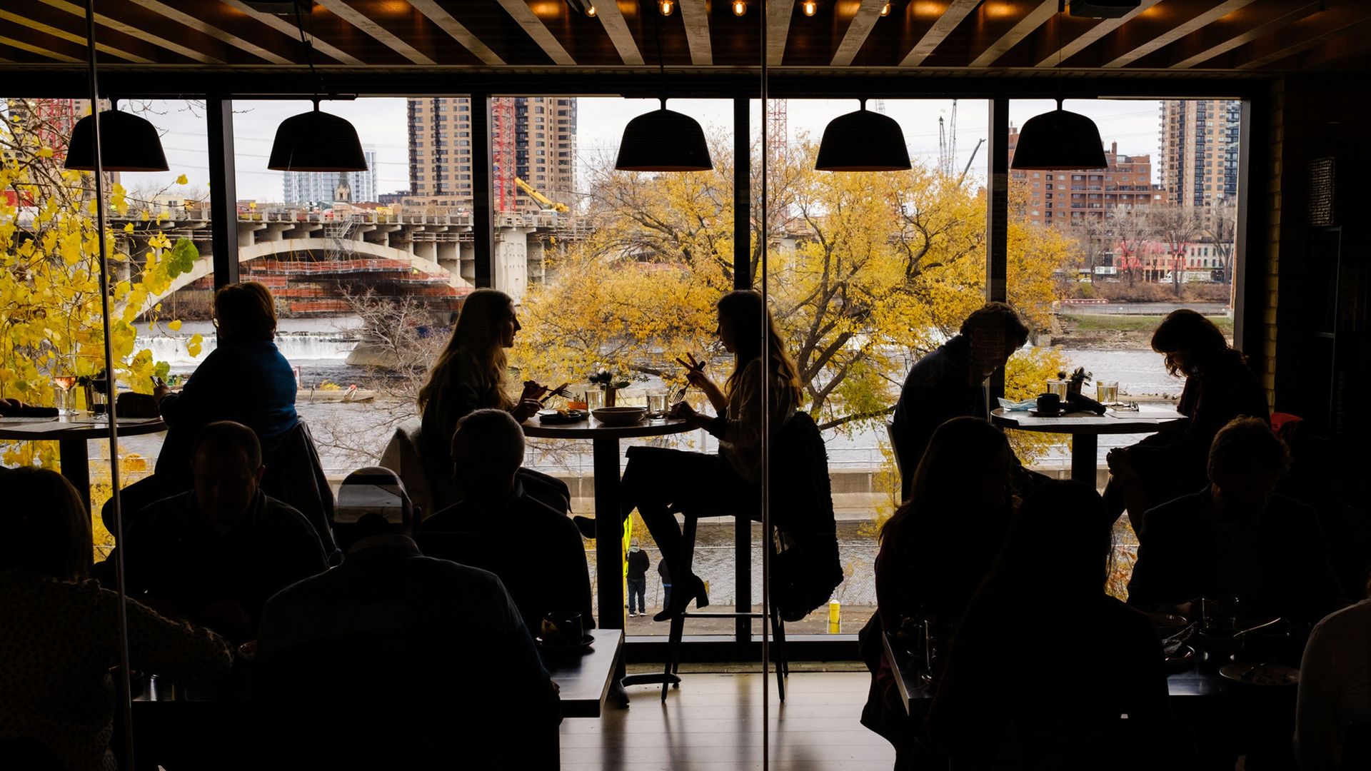 Silhouetted people dining inside a restaurant by large windows showing autumn trees with yellow leaves, a river, and city buildings and a bridge in the background.
