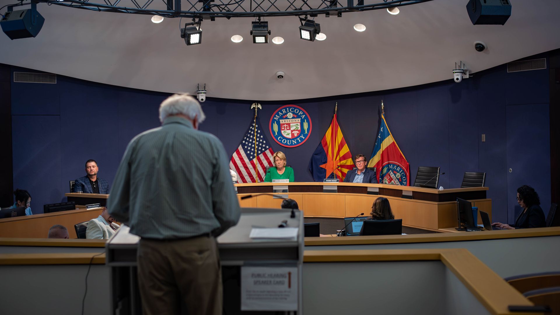 Panel of officials sits behind a curved desk in a conference room, flags (US, Arizona, county seal) visible; overhead stage lights and an audience seated in front.