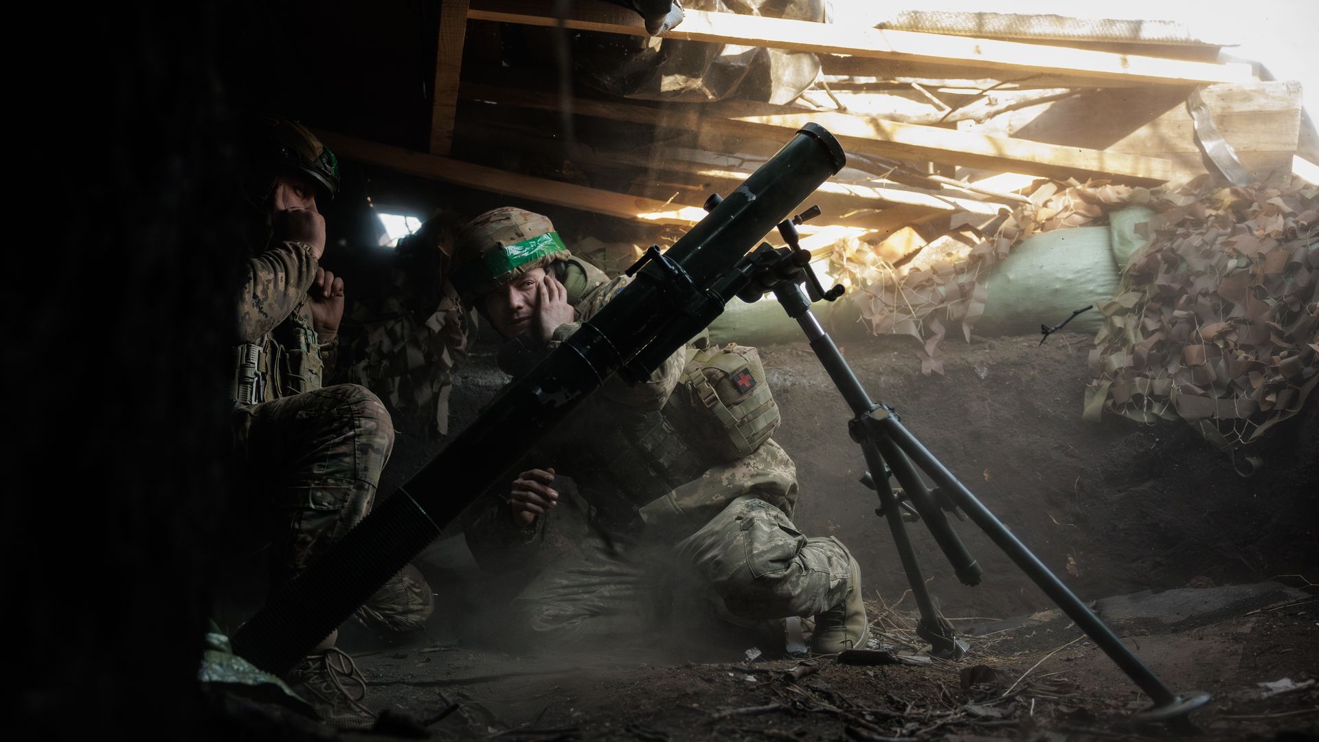 Two soldiers in camouflage gear and helmets crouch inside a dim dugout with wooden beams and camouflage netting, next to a large mounted mortar weapon.