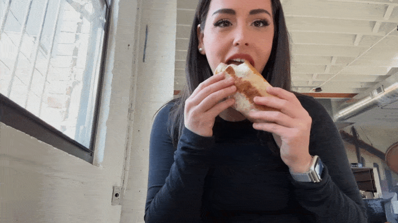 A woman with long dark hair in a black long-sleeve top sits indoors, biting into a crusty sandwich she holds with both hands, looking toward the camera near a window.
