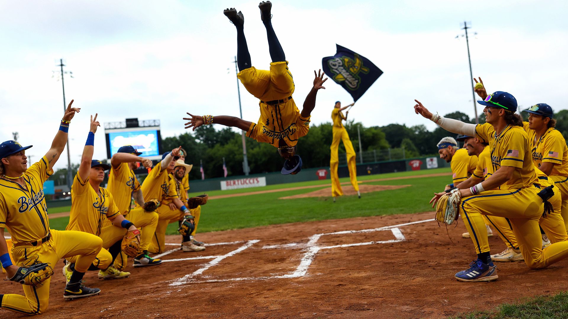 A Savannah Bananas player does a backflip next to kneeling teammates holding gloves. 