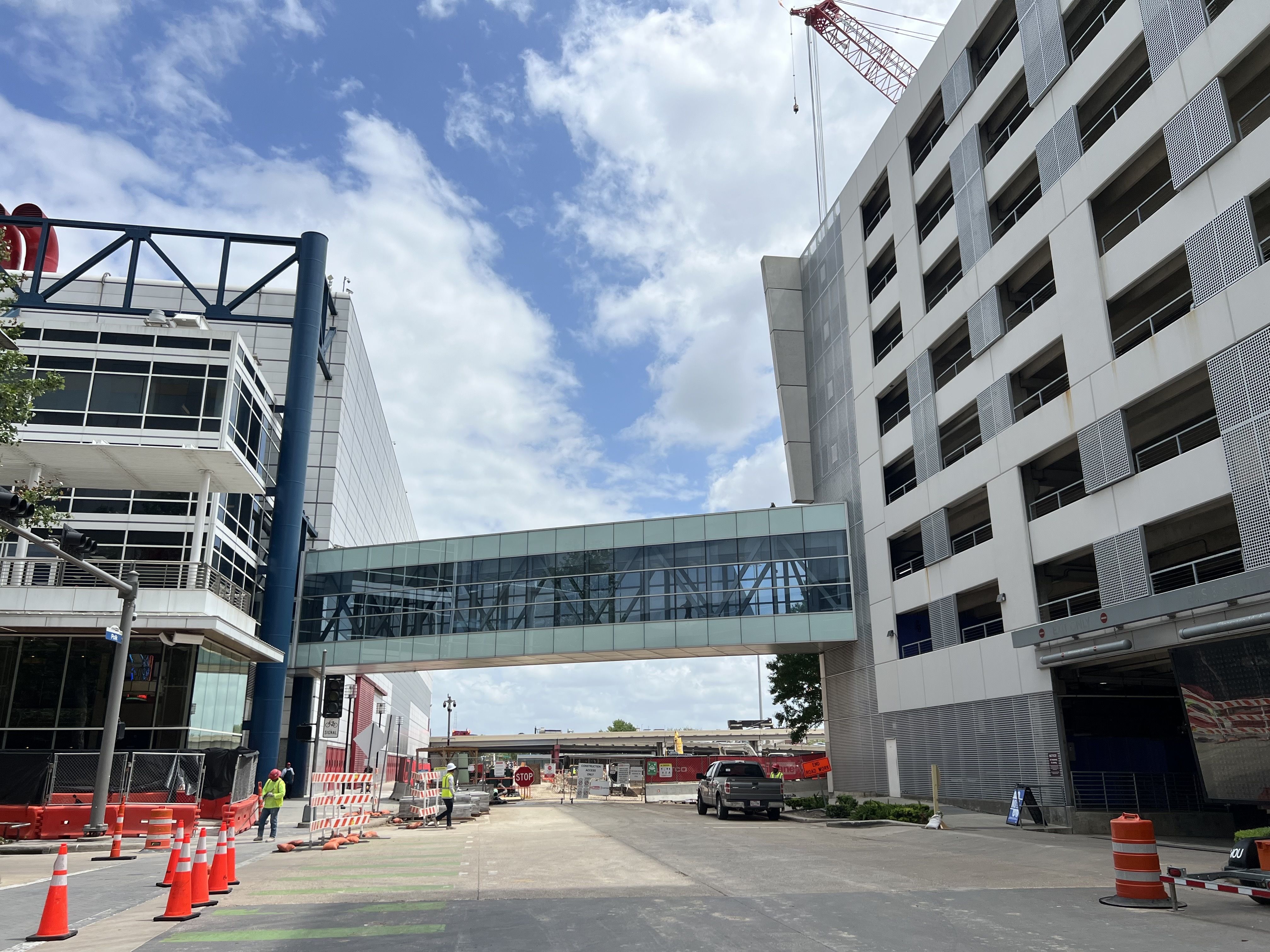 Urban construction site with two white buildings joined by a glass skybridge. A tall crane looms above; orange cones line the street. Workers in yellow vests near a parked SUV under a blue sky.