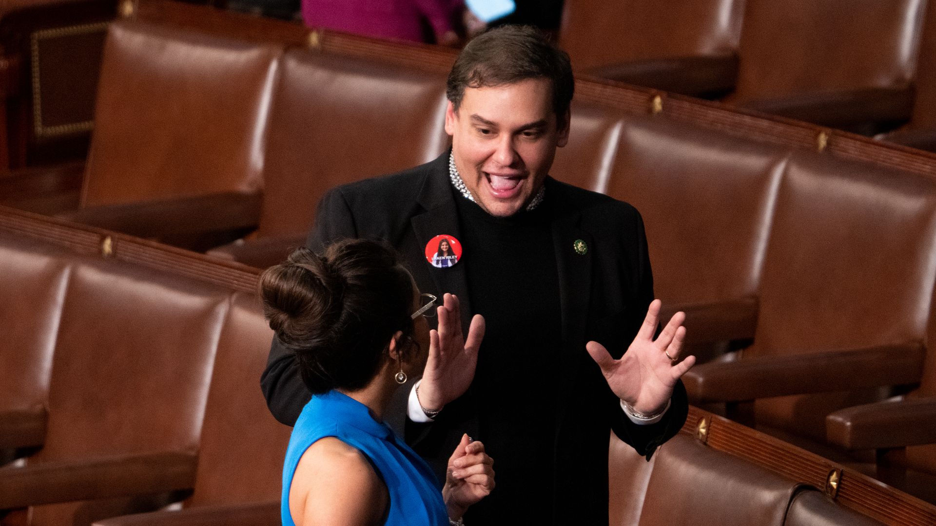 Former Rep. George Santos, wearing a dark gray suit over a black sweater with a red button, standing on the House floor talking to Rep. Lauren Boebert, wearing a blue dress.