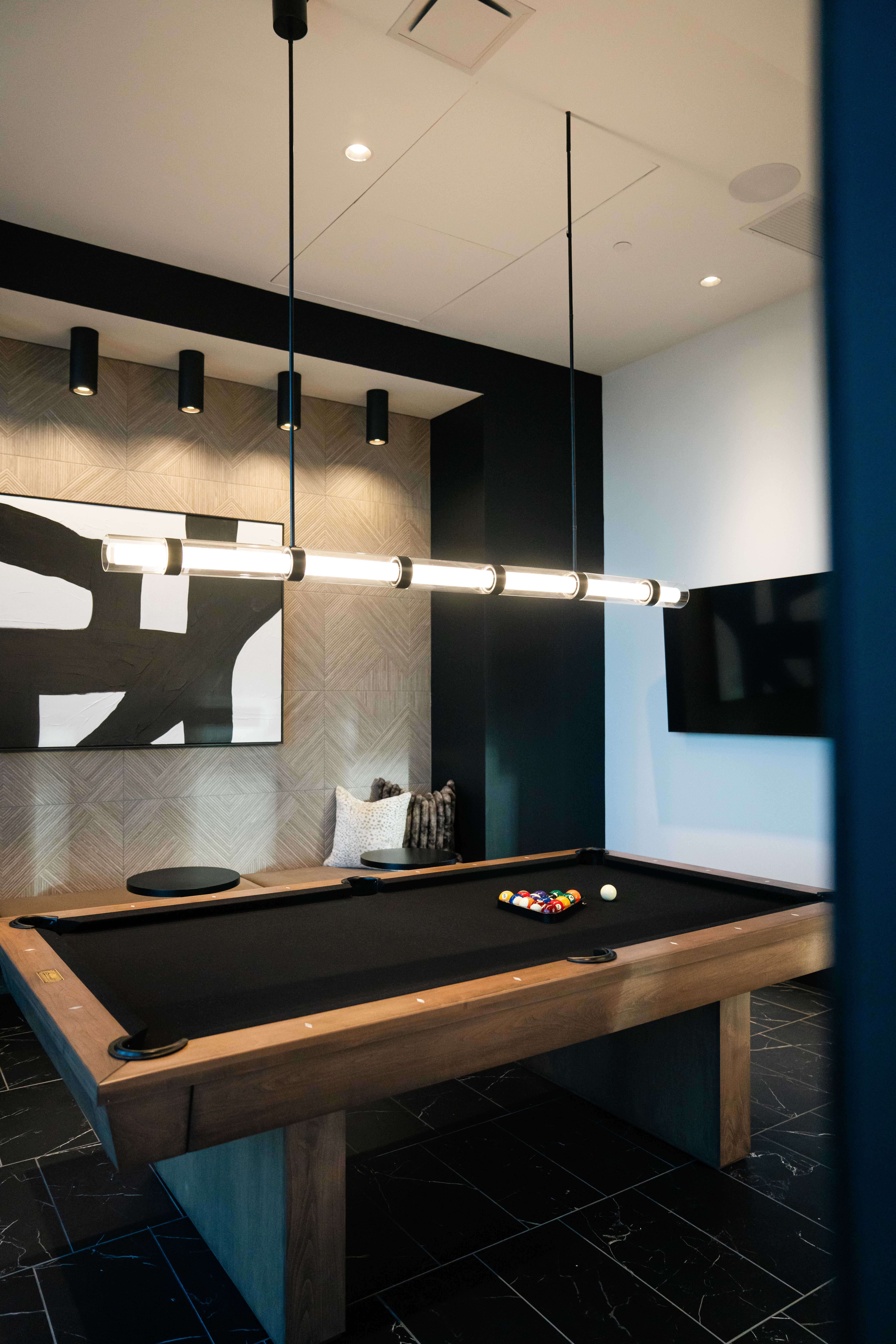 Modern game room with a black felt pool table, wooden frame, unique linear pendant light above, black and beige walls, abstract black and white wall art, and a flat-screen TV.