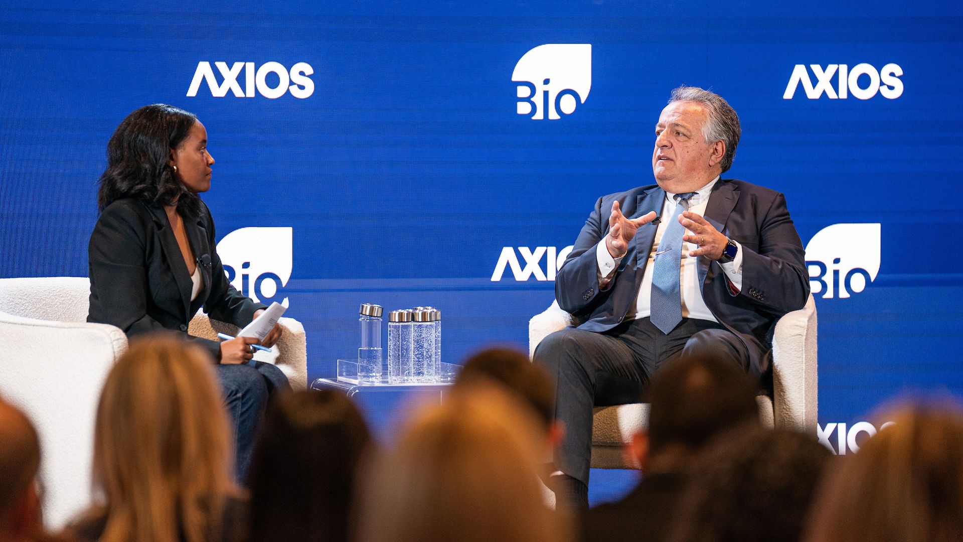 Panel on a blue Axios backdrop: a man in a suit speaks from the right, a woman interviewer listens on the left, with a blurred audience and water bottles on a table.