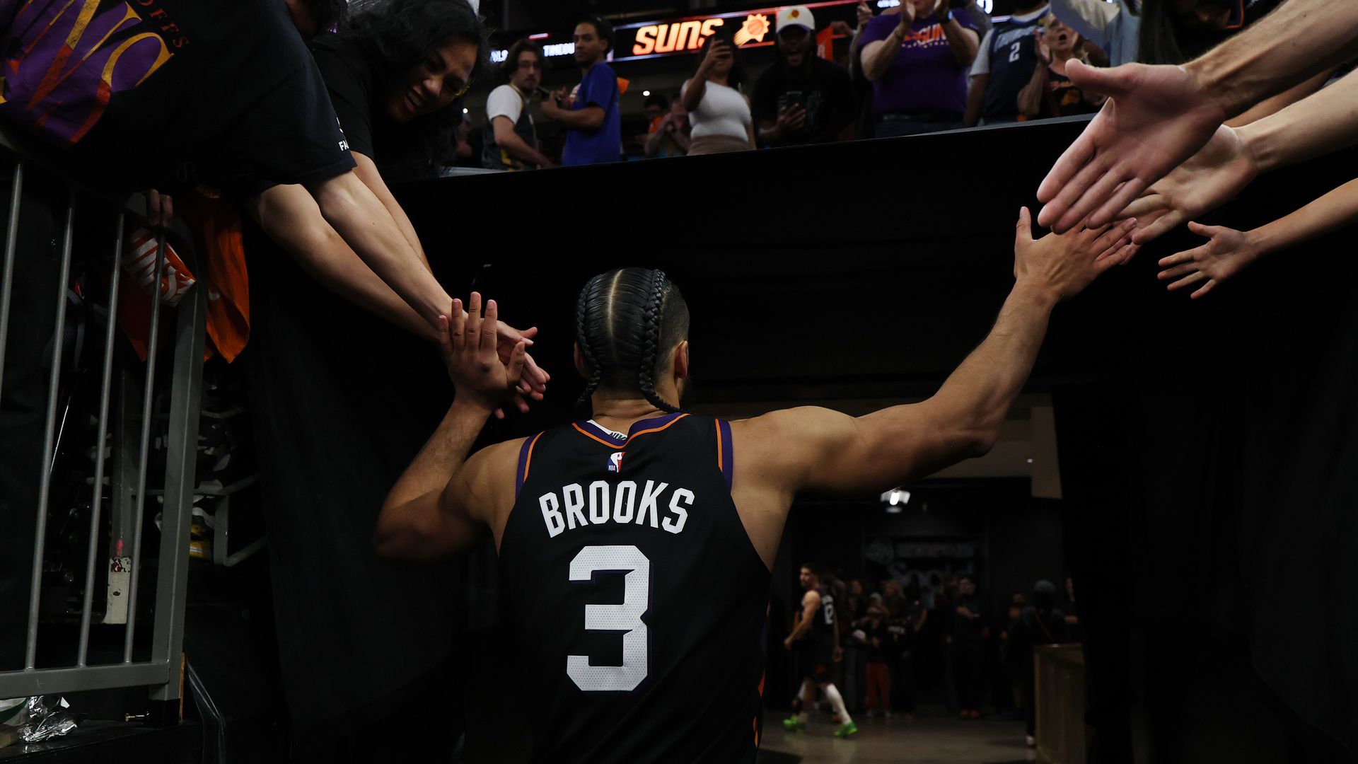 A basketball player in a black jersey, viewed from behind, with the word "Brooks" and the number "3" on the back of his uniform, slaps hands with fans on both sides of him as he walks off the court and into the tunnel.