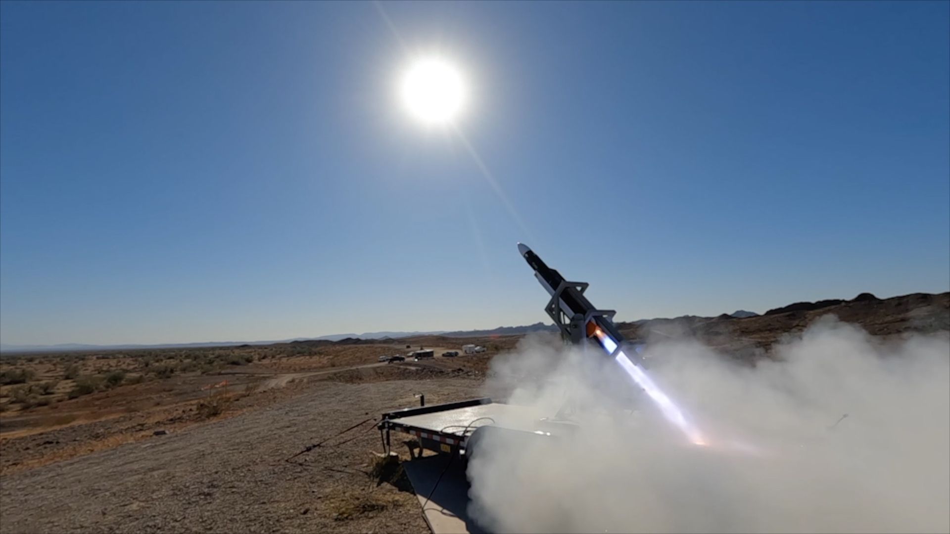Rocket launching from a desert platform with bright sun overhead, clear blue sky, and smoke billowing around the launch site.