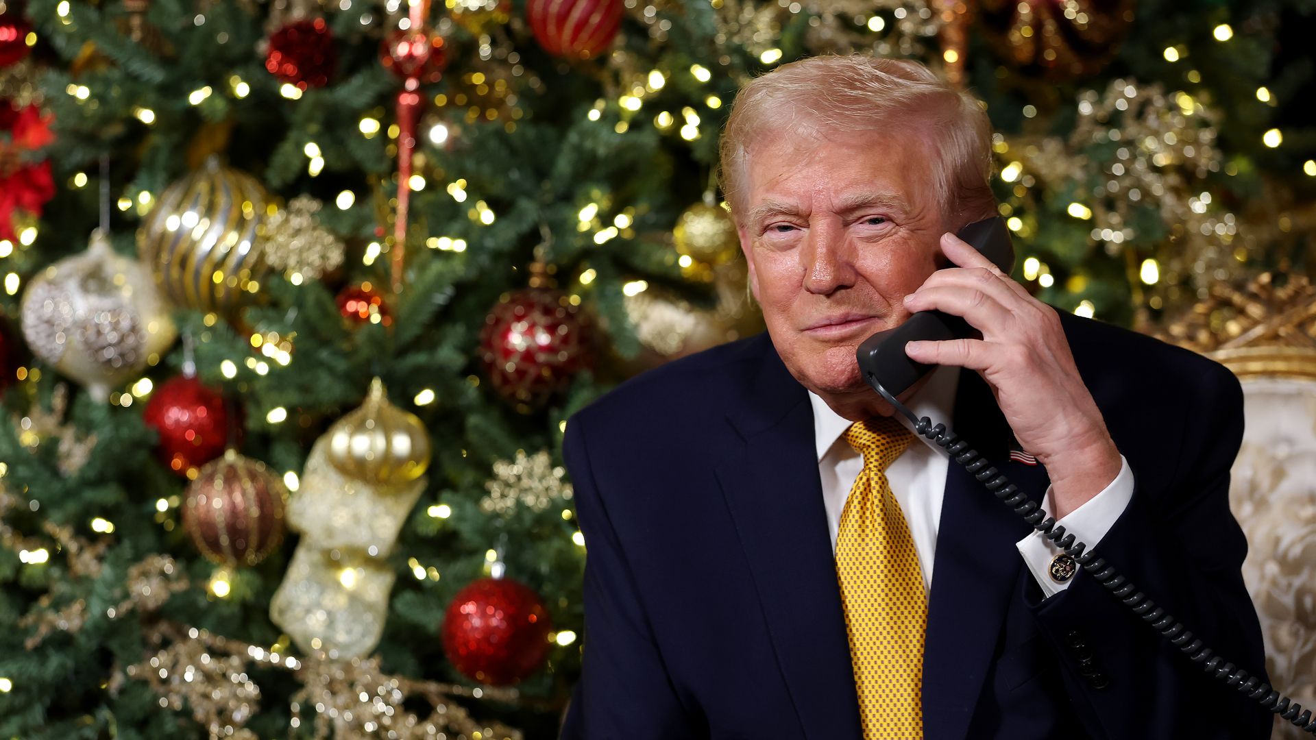 A fair, tan-skinned, graying President Trump, wearing a navy suit jacket, white shirt and gold tie with black polka dots on it smiles as he holds a 1980s-style black telephone with a coil cord while sitting on a chair with gold trim in front of a heavily decorated Christmas tree.