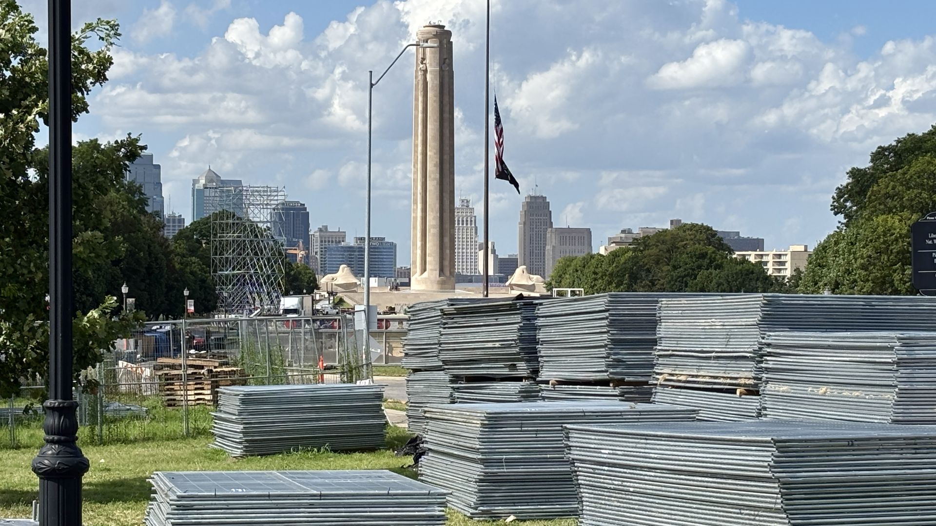 Construction site in a city park: foreground stacks of metal sheets, a tall stone obelisk with an American flag, city skyline and blue sky with clouds behind, green trees on both sides.