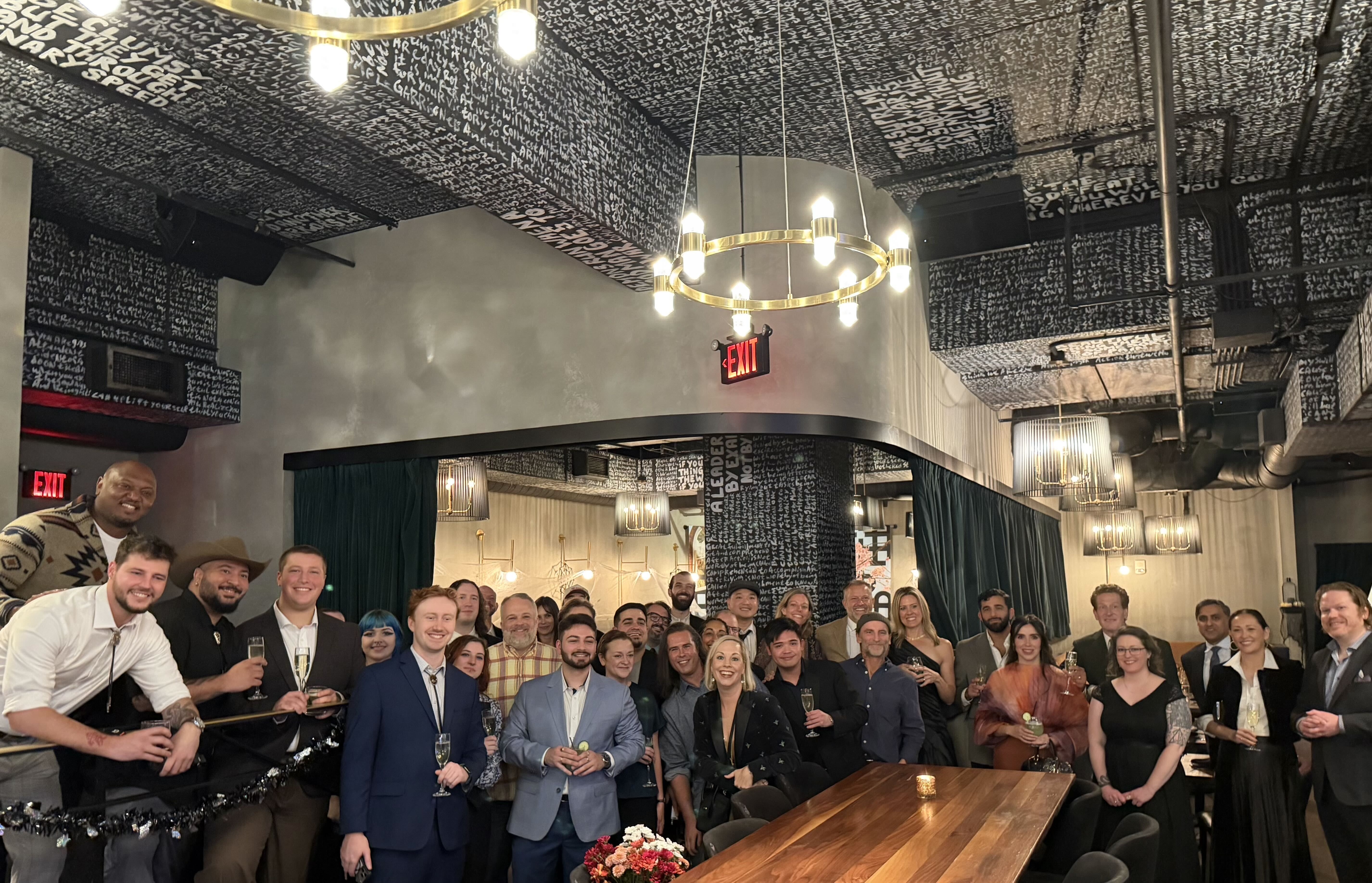 Large group of well-dressed people posing in a bar/restaurant with a black ceiling covered in white handwritten text, gold circular chandeliers, and an EXIT sign in the background.