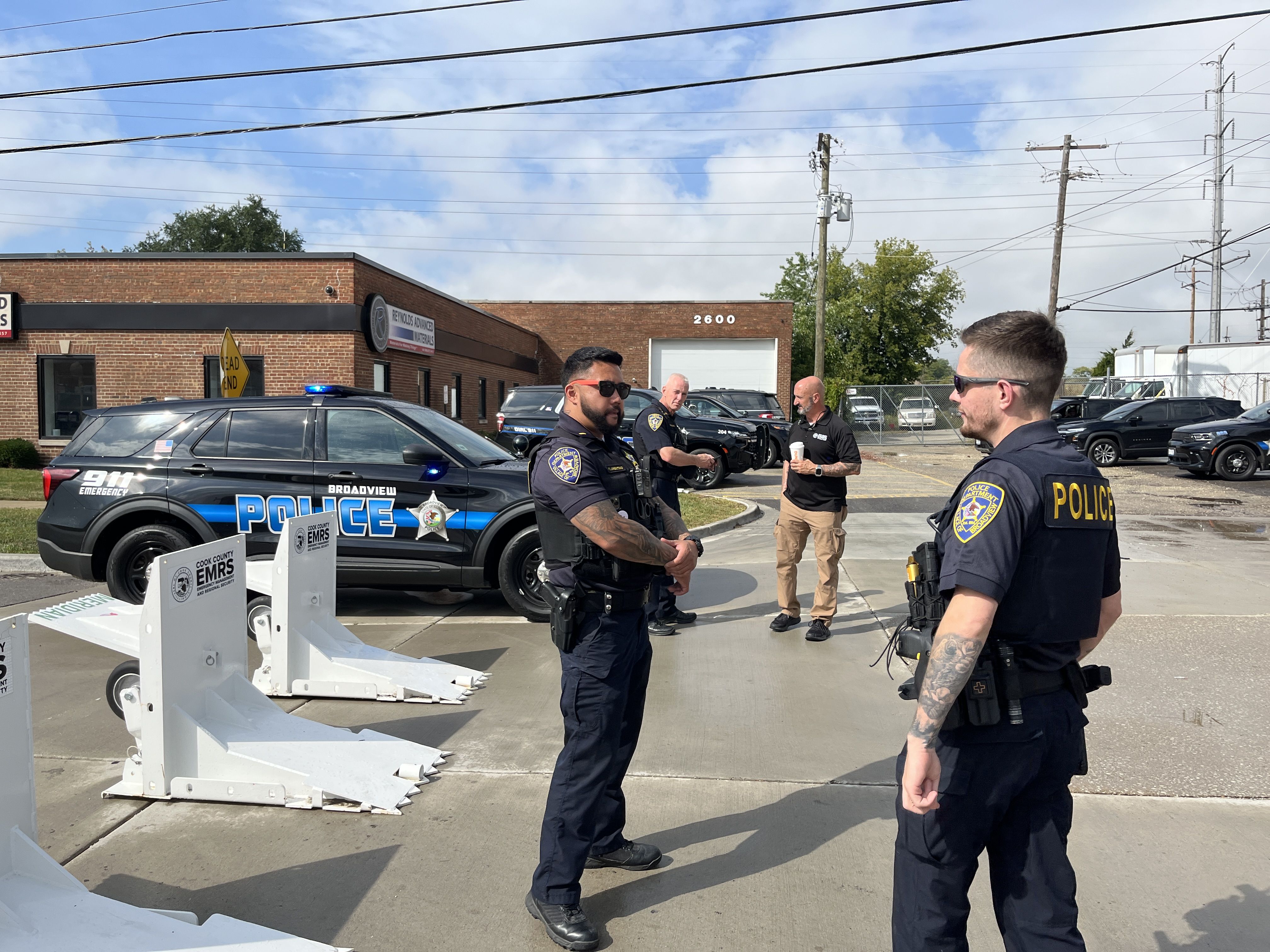 Four police officers in black uniforms with blue and yellow police patches stand and talk near black and blue police SUVs outside a brick building with the address 2600 under a blue sky.