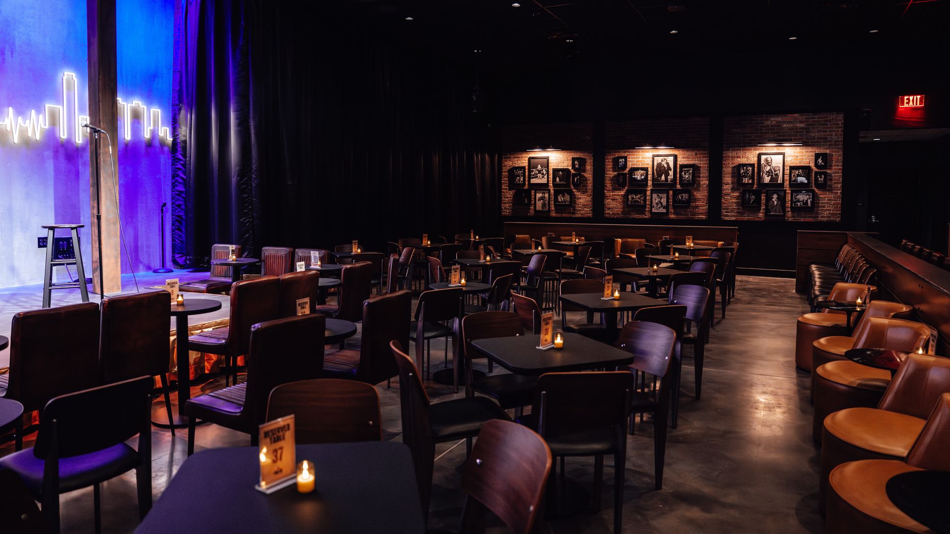 Dimly lit comedy club with empty tables and chairs, a small stage with microphone and stool, blue and purple lighting, and brick walls featuring black-and-white framed photos.