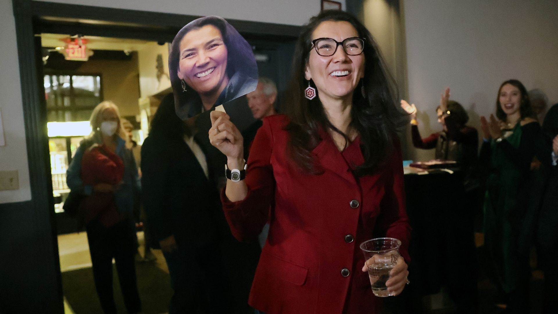 Rep. Mary Peltola, wearing a red pant suit and holding a glass of water and a cutout of her own face.