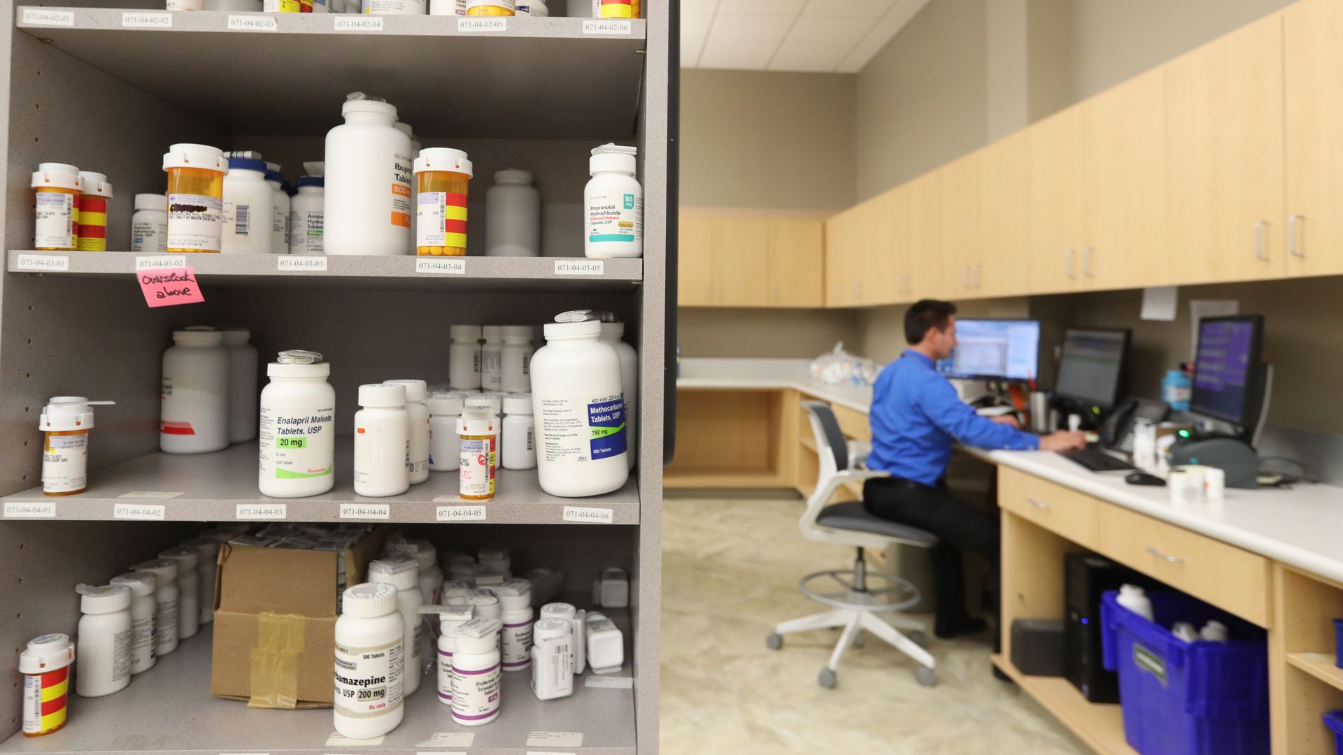 A pharmacy technician works at a desk with drugs sitting on a shelf in the foreground.